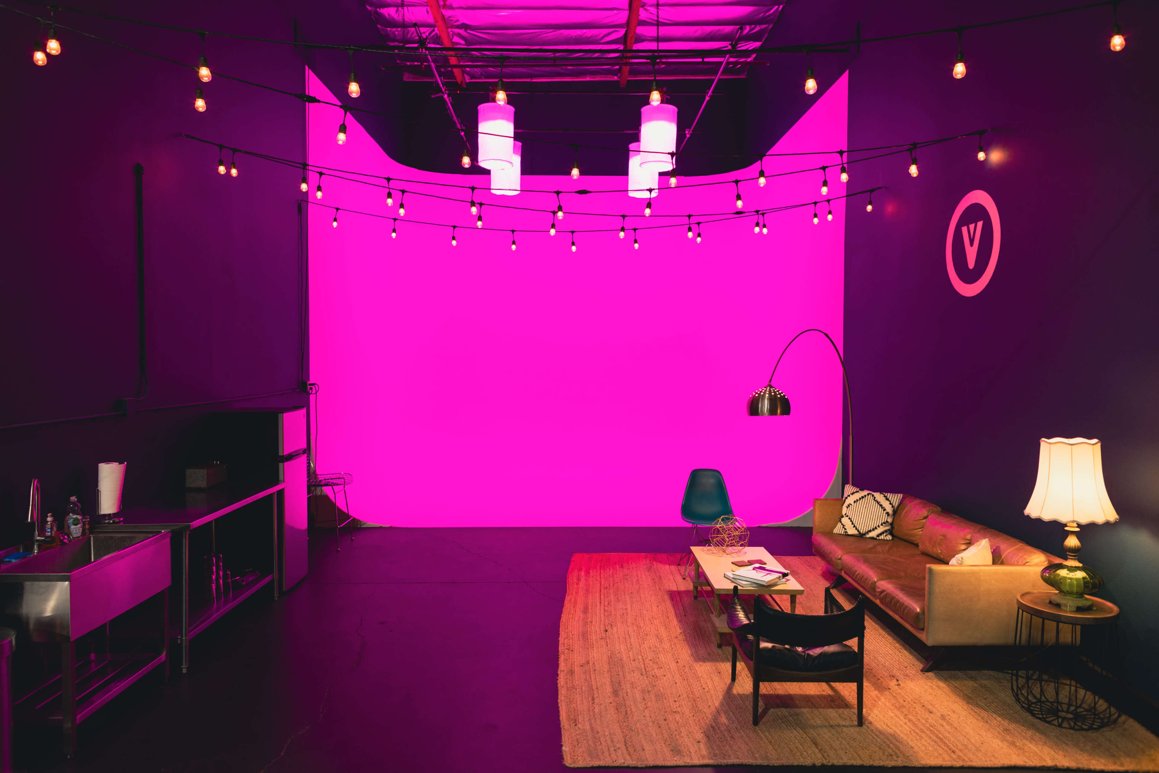A modern lounge area features a sofa, a coffee table, and a lamp against a vibrant pink backdrop, with string lights illuminating the space.