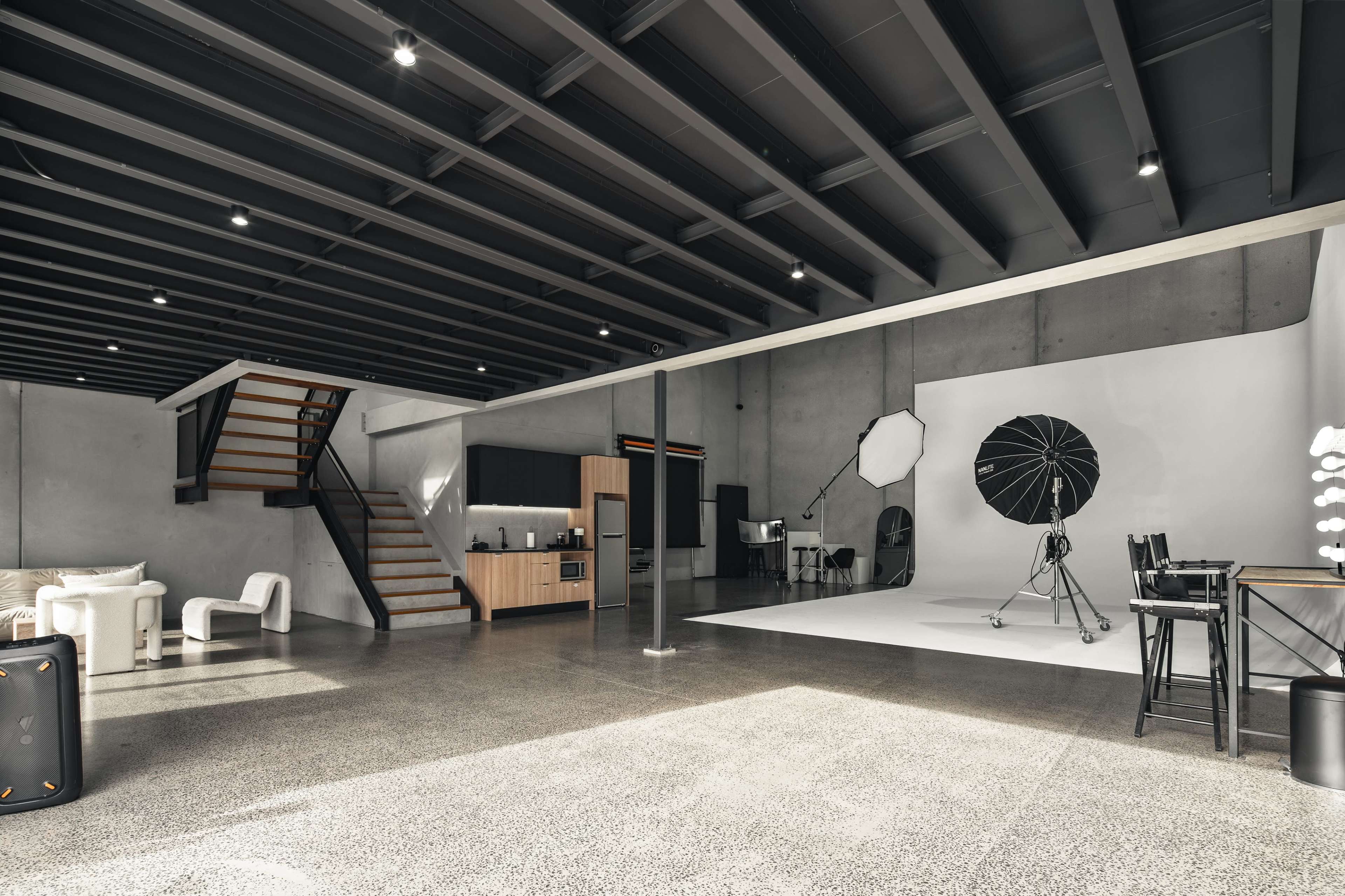 The image shows a spacious interior featuring a kitchen area, a living space with modern furniture, a staircase, and a photography studio setup with a backdrop and lighting equipment.