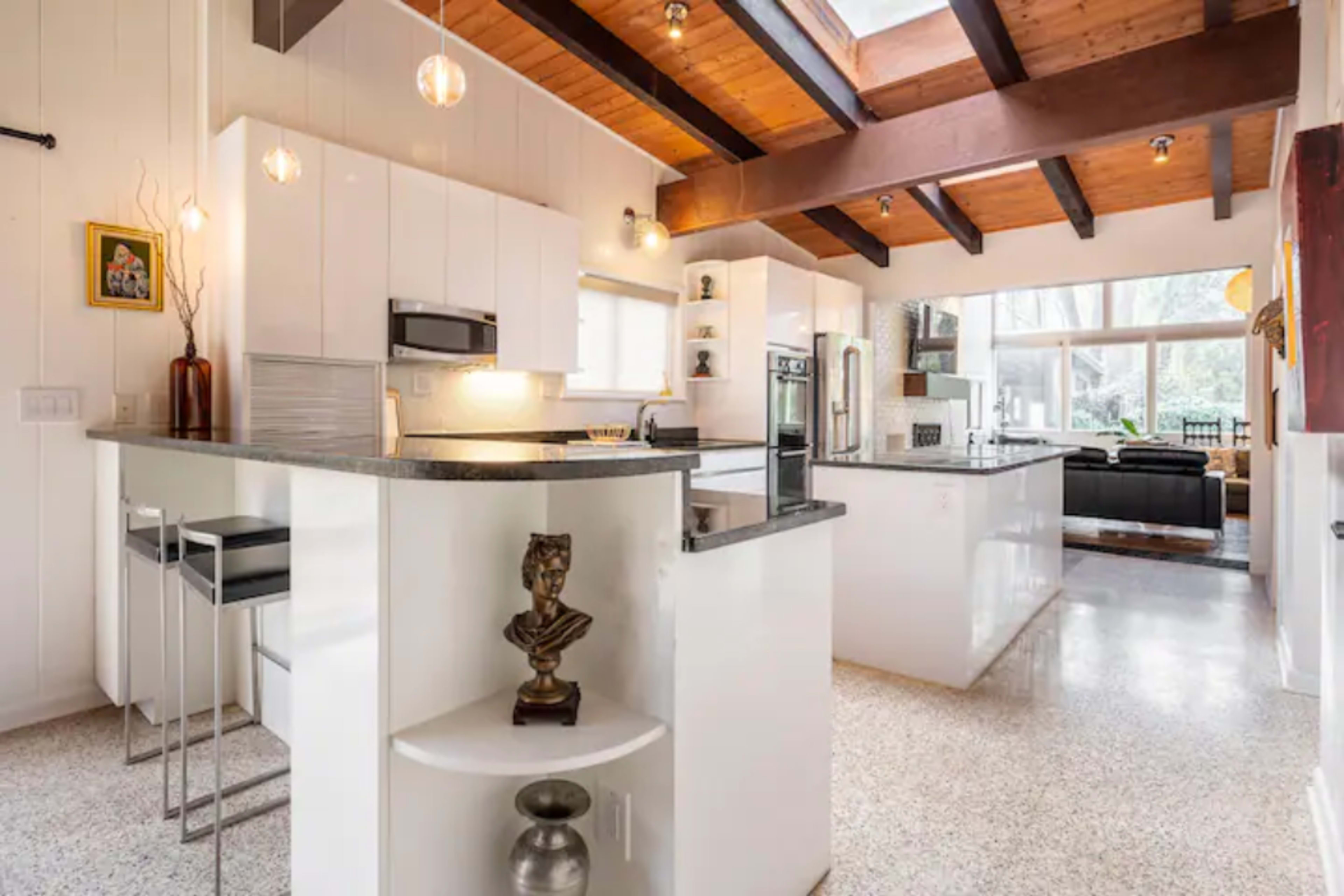 The image shows a modern kitchen with white cabinets, a granite countertop, and a spacious open layout that connects to a living area with large windows.