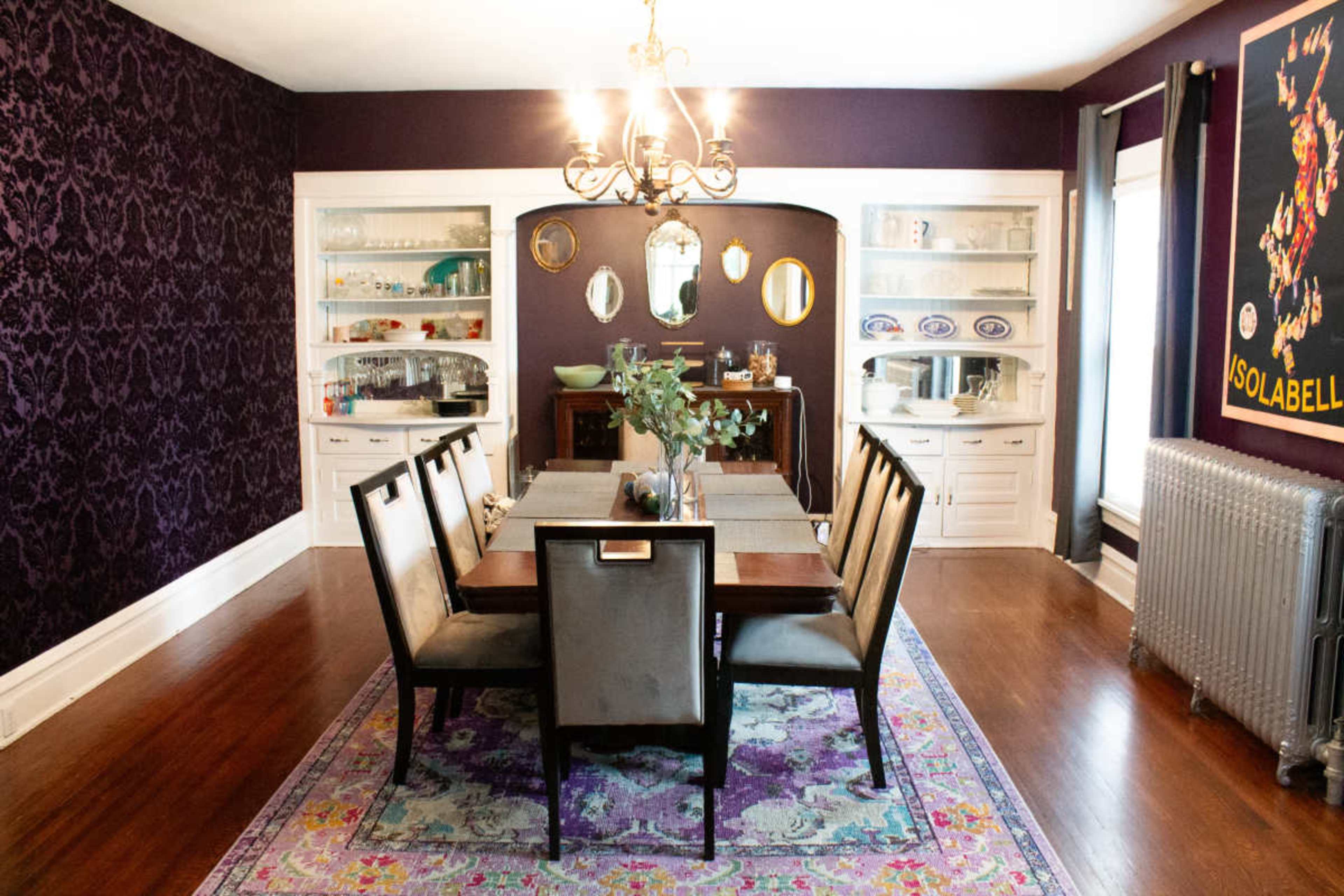 The dining room features a long wooden table surrounded by upholstered chairs, with a patterned rug, wall-mounted shelves displaying glassware, and purple wallpaper.