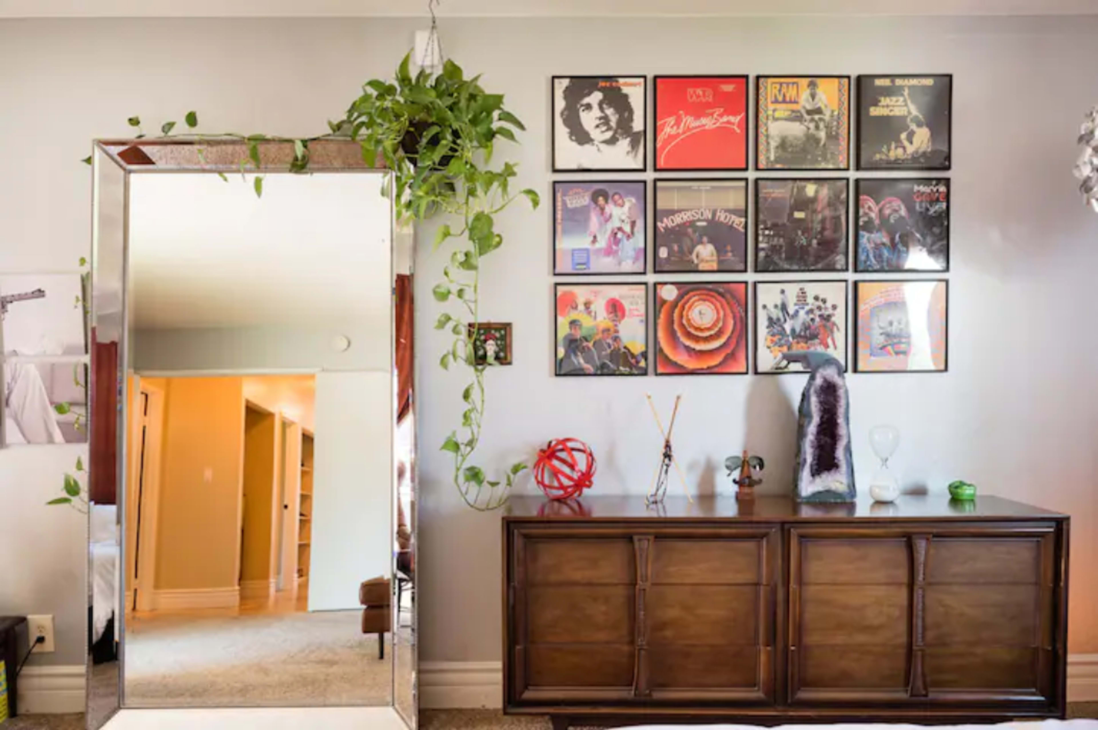A wall adorned with framed vinyl records above a wooden sideboard, with a large mirror reflecting a room in the background and plants hanging decoratively.