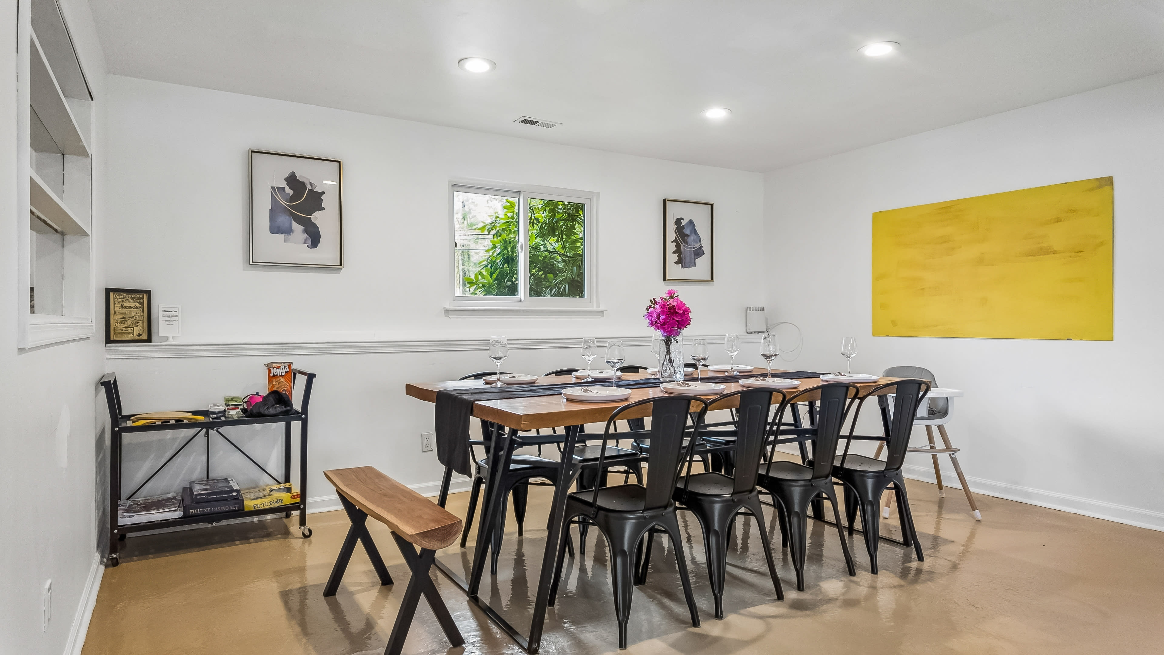 A dining area features a large wooden table with black chairs, set for a meal, surrounded by minimalist decor and natural light from a window.