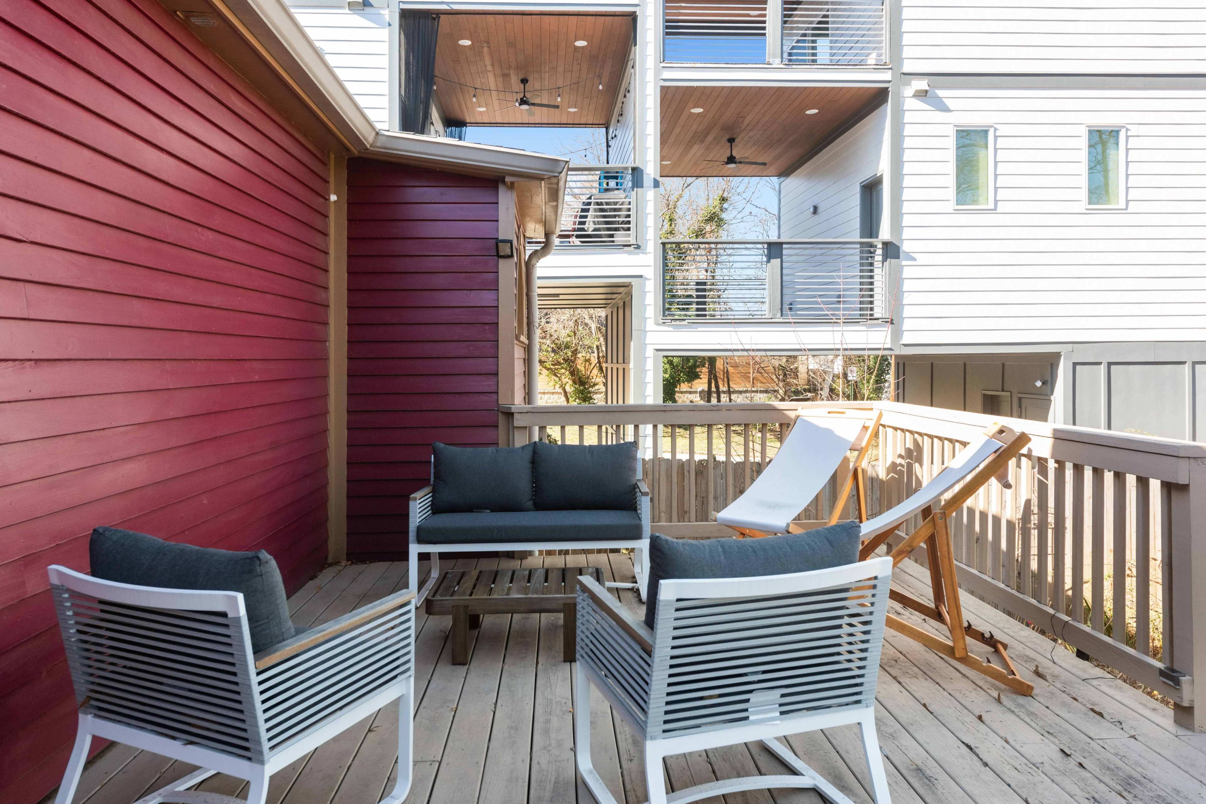 The image shows a wooden deck with white outdoor furniture, including two chairs and a table, set against a backdrop of a red wall and a modern building with multiple balconies.