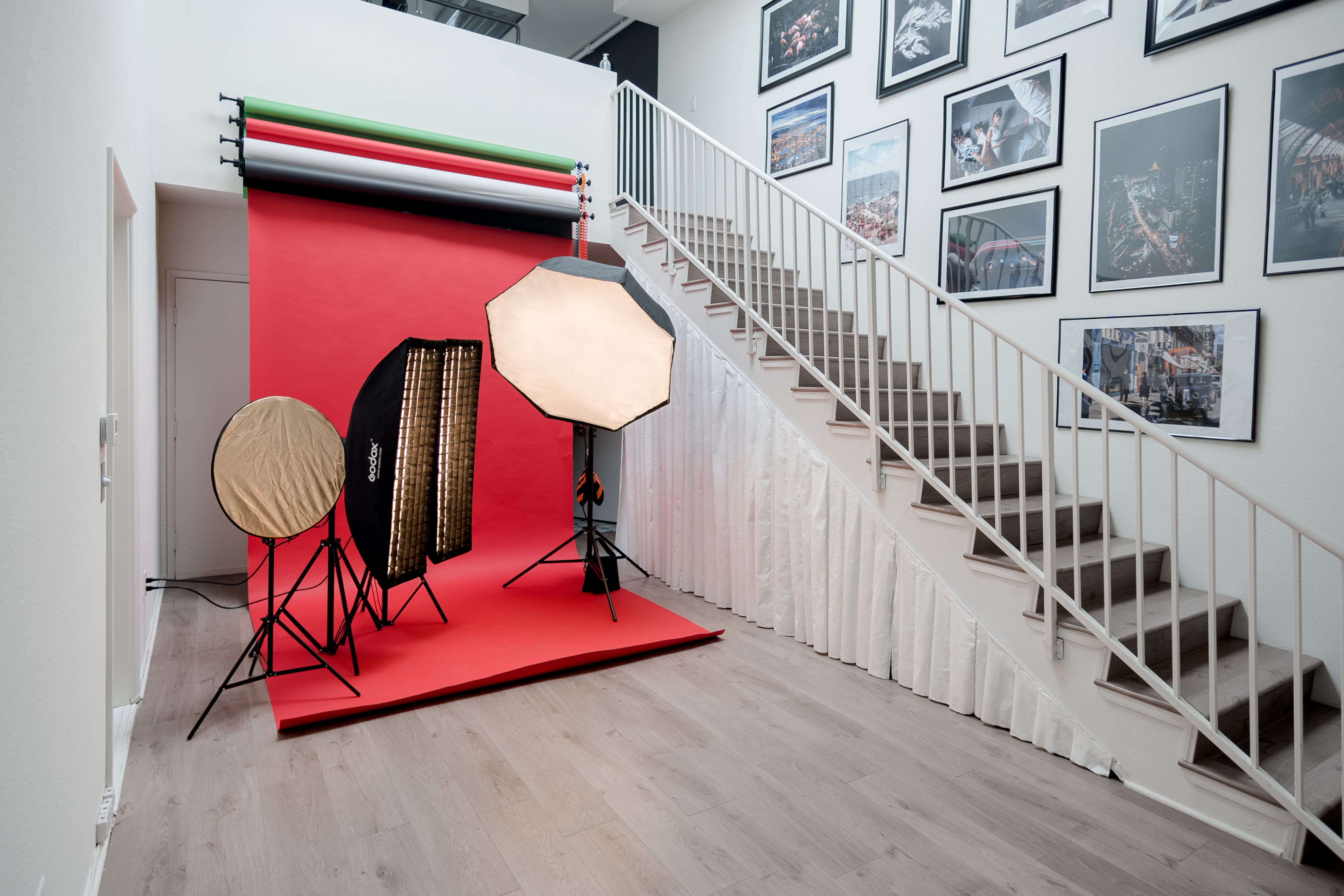 A photography studio setup with lighting equipment positioned against a red backdrop, located next to a staircase lined with framed photographs on the wall.