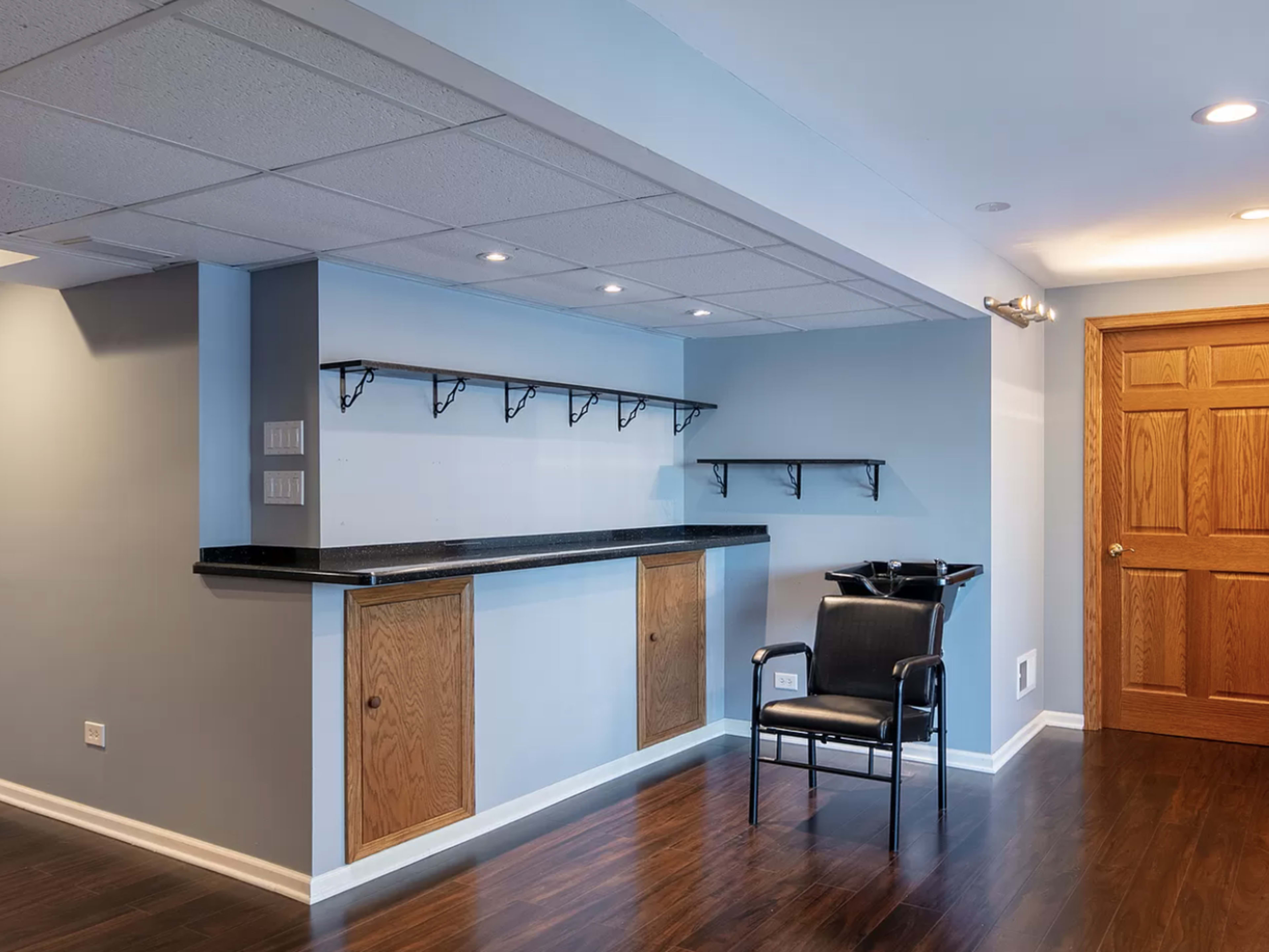 The image shows a sparsely furnished room with a black chair, a long countertop, shelving above, and wooden doors.