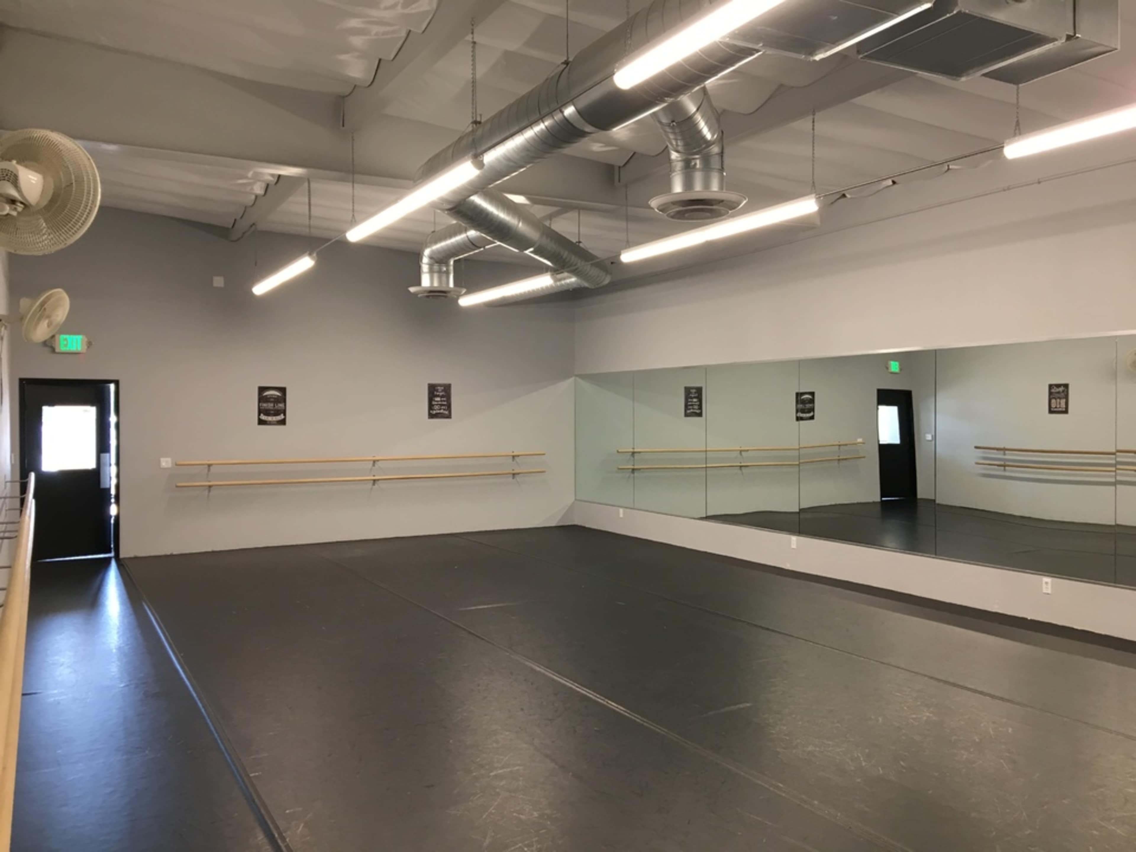 The image shows an empty dance studio with a gray floor, wall-mounted barres, large mirrors, and fluorescent lighting.
