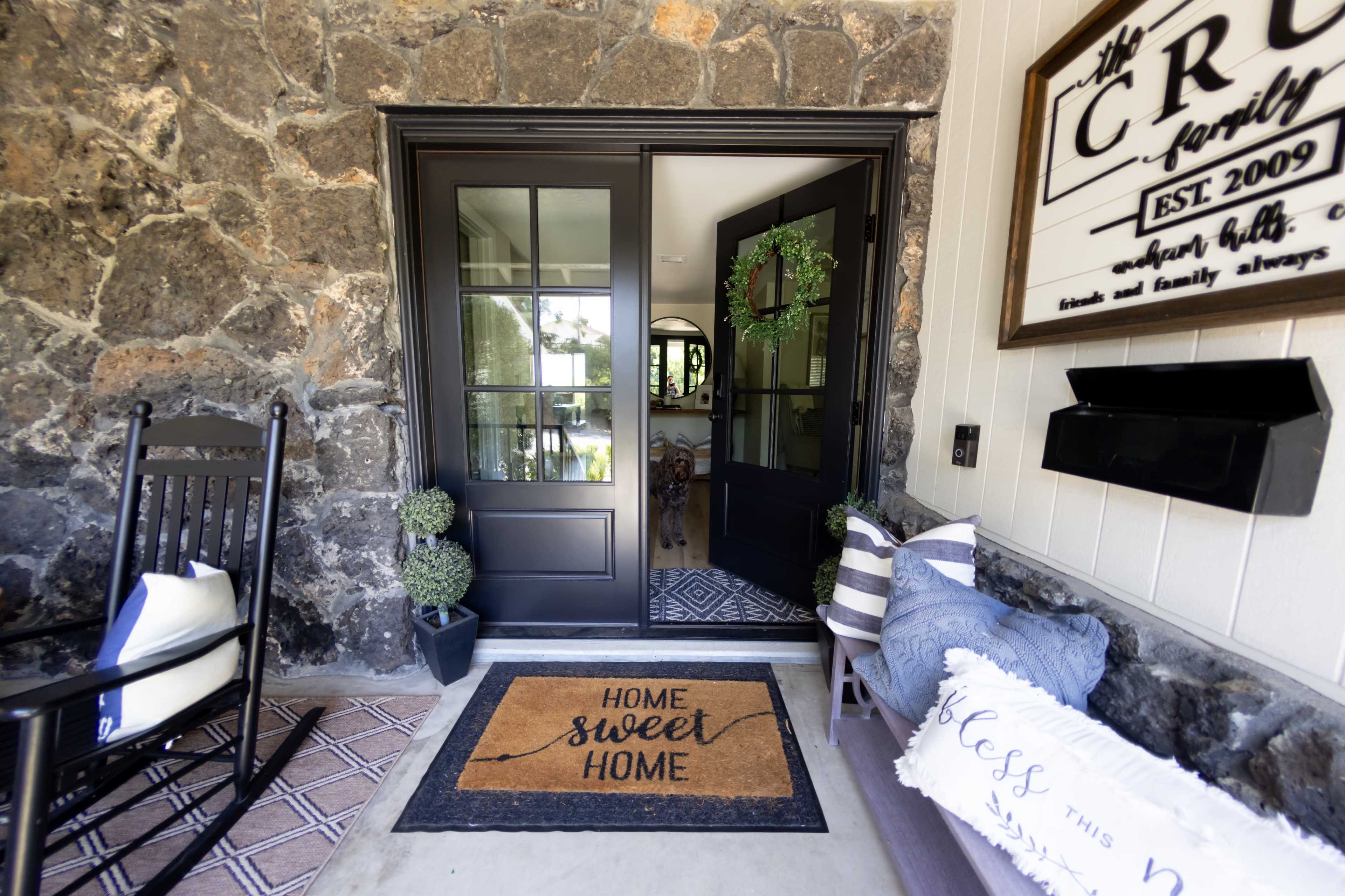 A stone-walled entrance with double black doors, a welcome mat reading "HOME sweet HOME," and decorative potted plants beside a rocking chair.