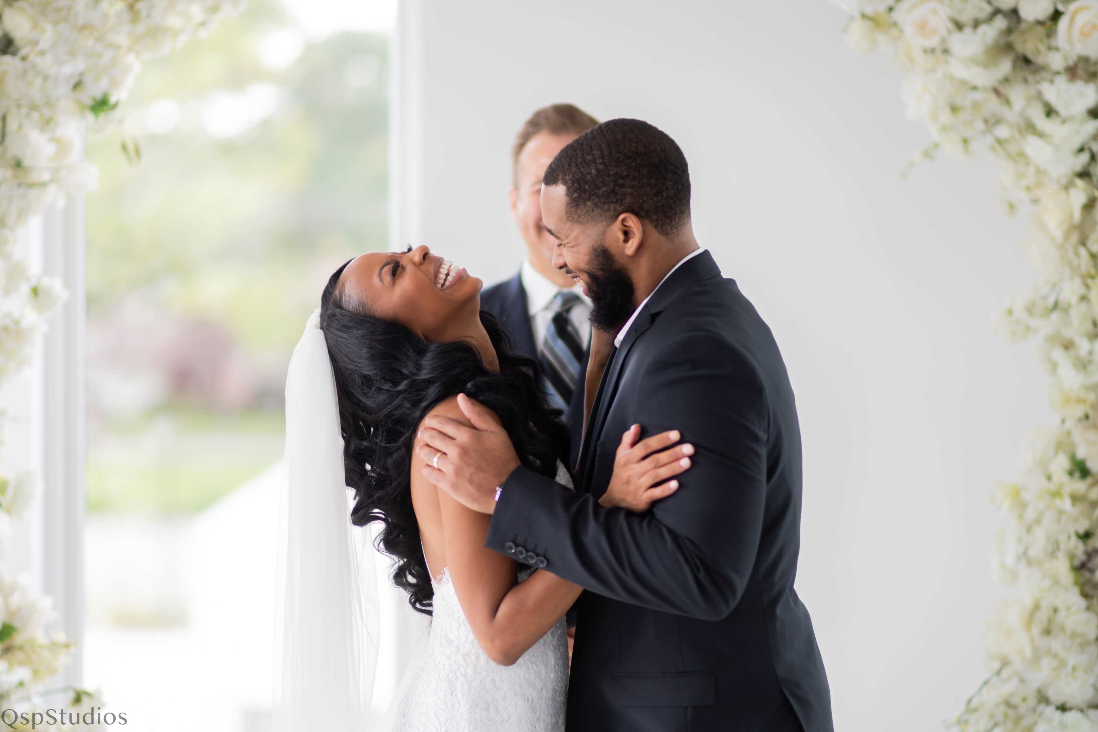 A couple smiles and embraces during their wedding ceremony, with a officiant standing behind them.