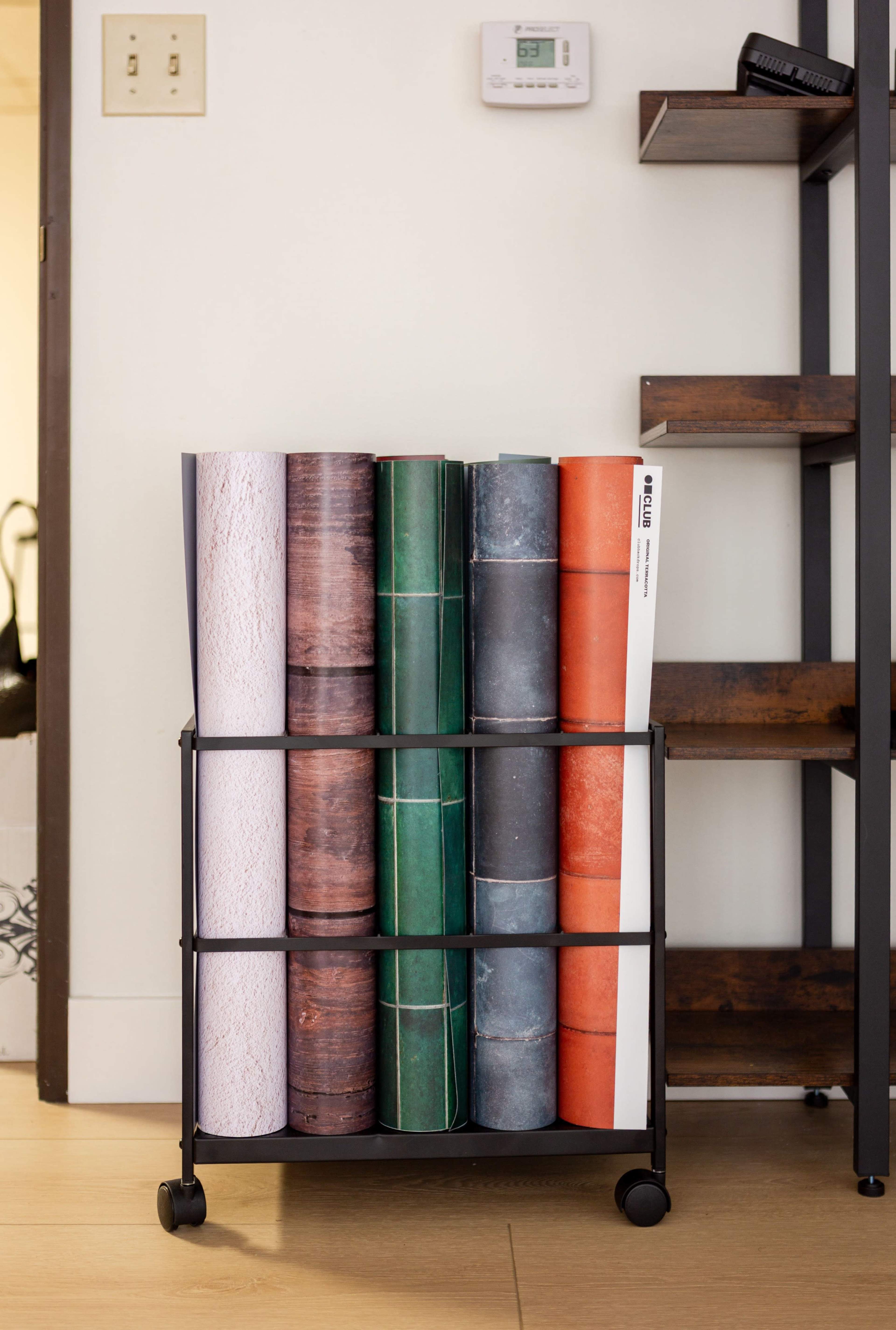 A metal cart holds several rolled papers in various colors and textures against a wall near a shelf.