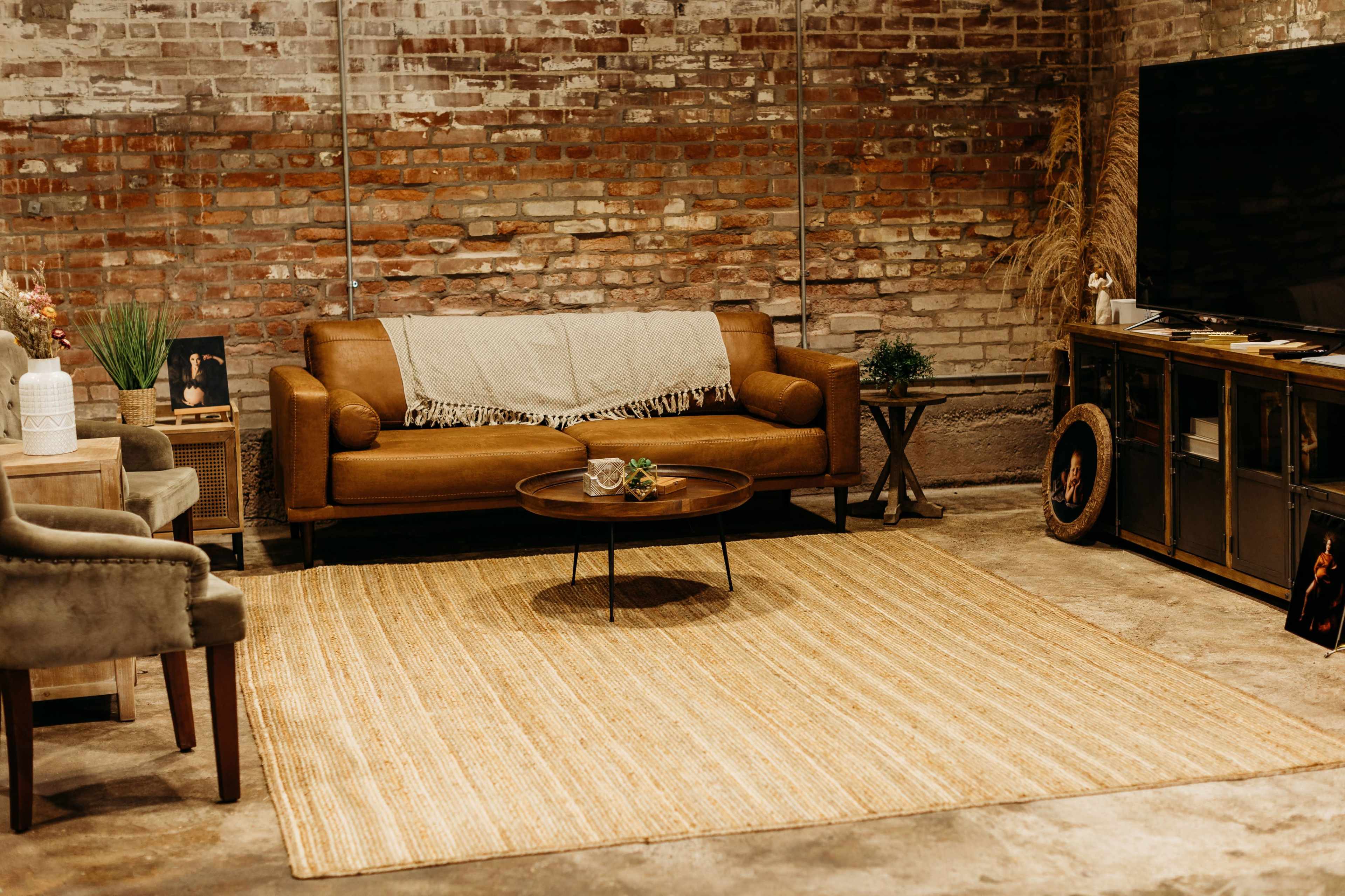 The image shows a cozy living room featuring a brown leather sofa, a round coffee table, a jute rug, and a textured brick wall.