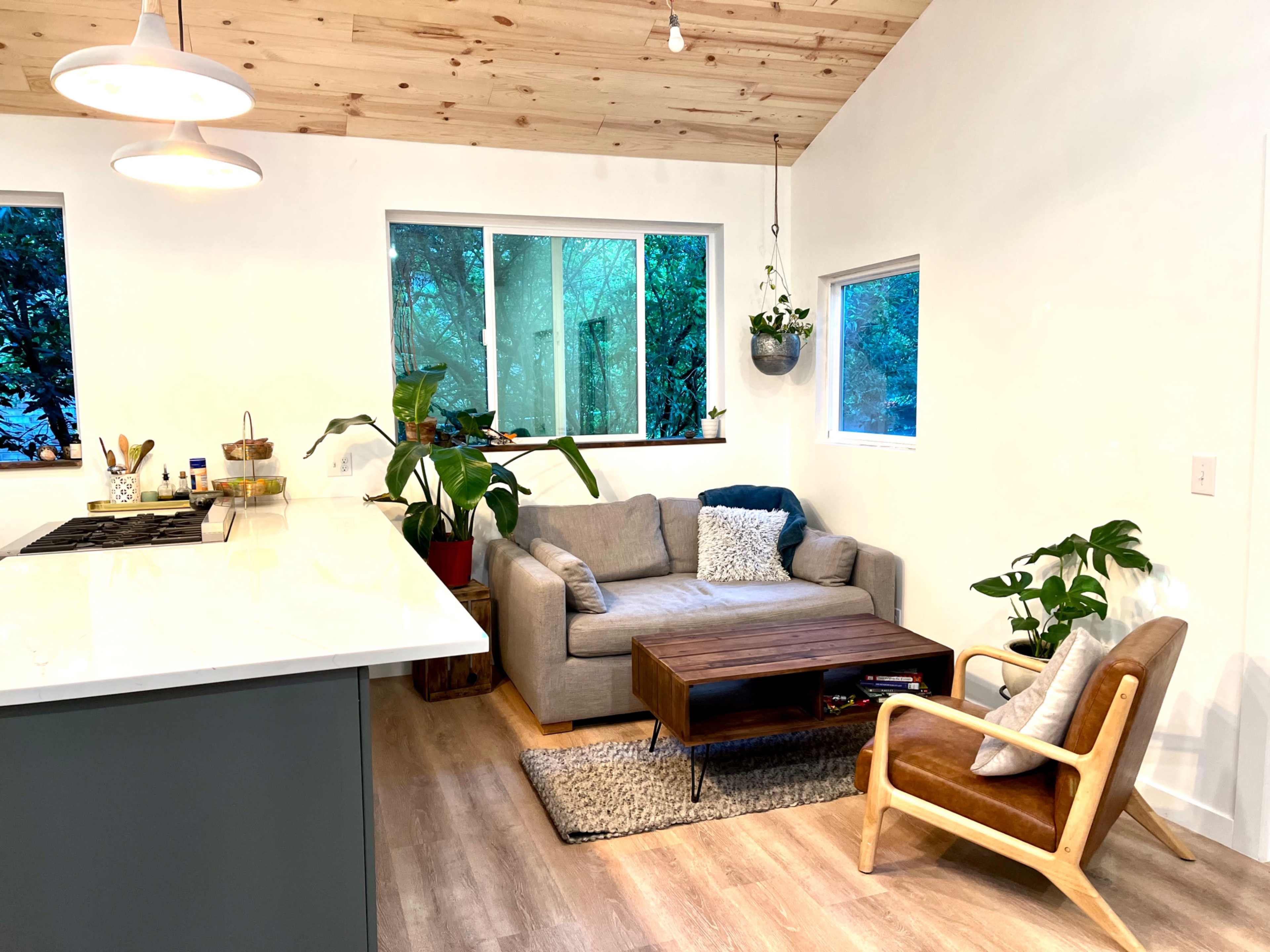 A modern living area featuring a gray sofa, a wooden coffee table, and plants, with large windows allowing natural light to enter.