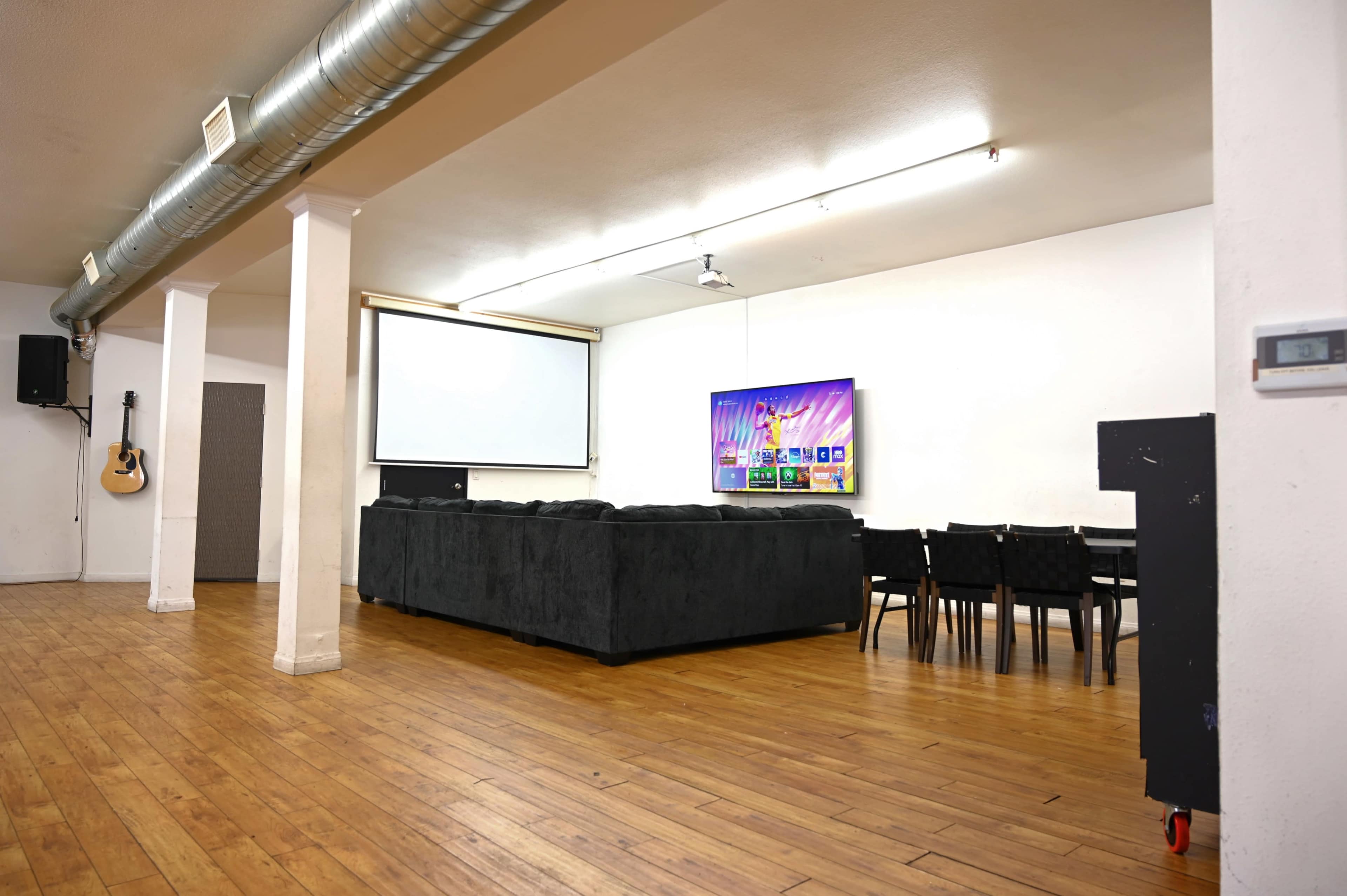A spacious room features a large sectional couch, a dining table with chairs, a projector screen, and a wall-mounted television.