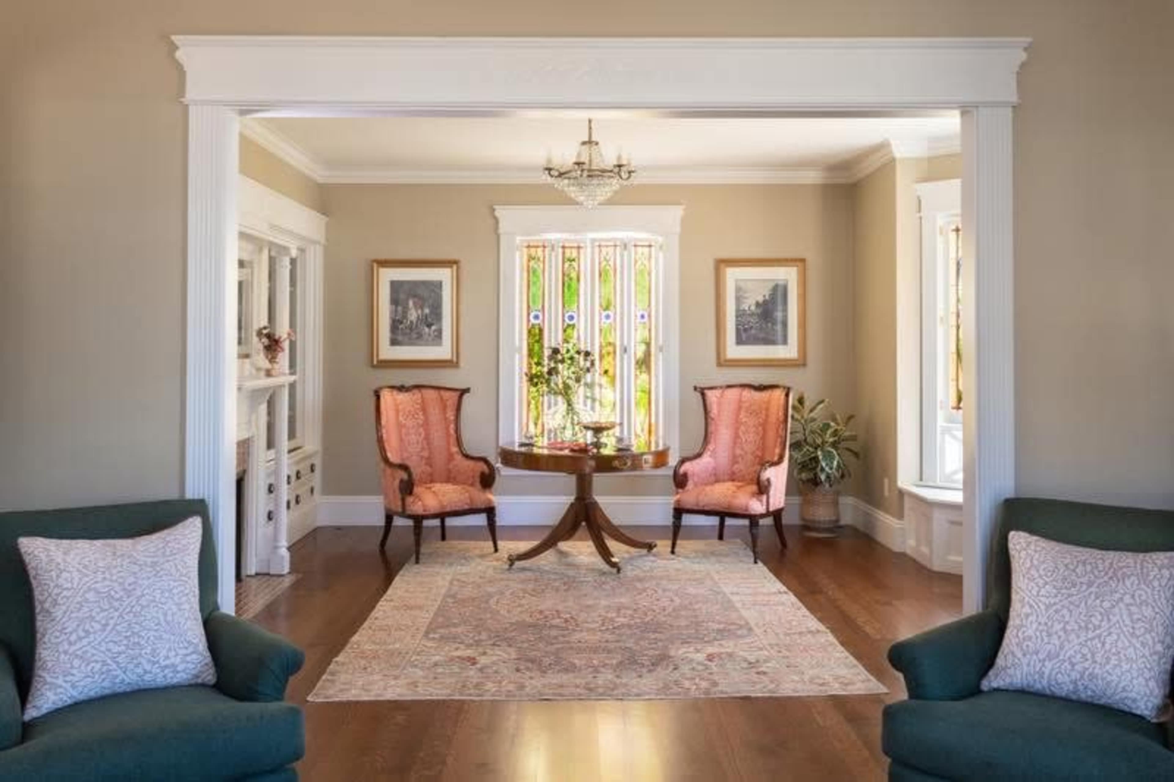 The image shows a well-lit living room featuring two orange armchairs and a circular wooden table, framed by ornate white moldings and a window with colorful drapes.