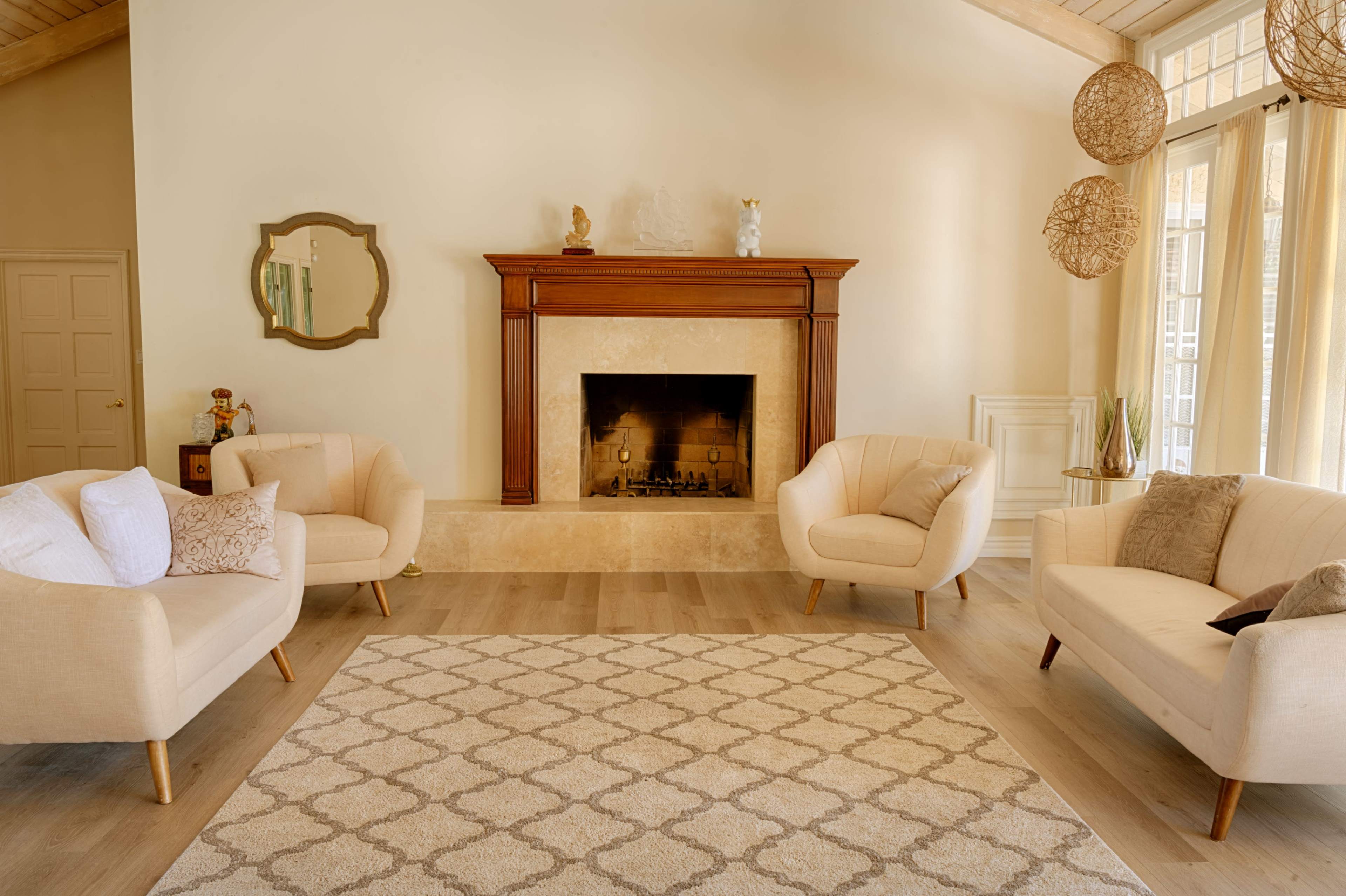 A spacious living room features two cream-colored armchairs and a matching sofa arranged around a patterned rug, with a stone fireplace and large windows in the background.