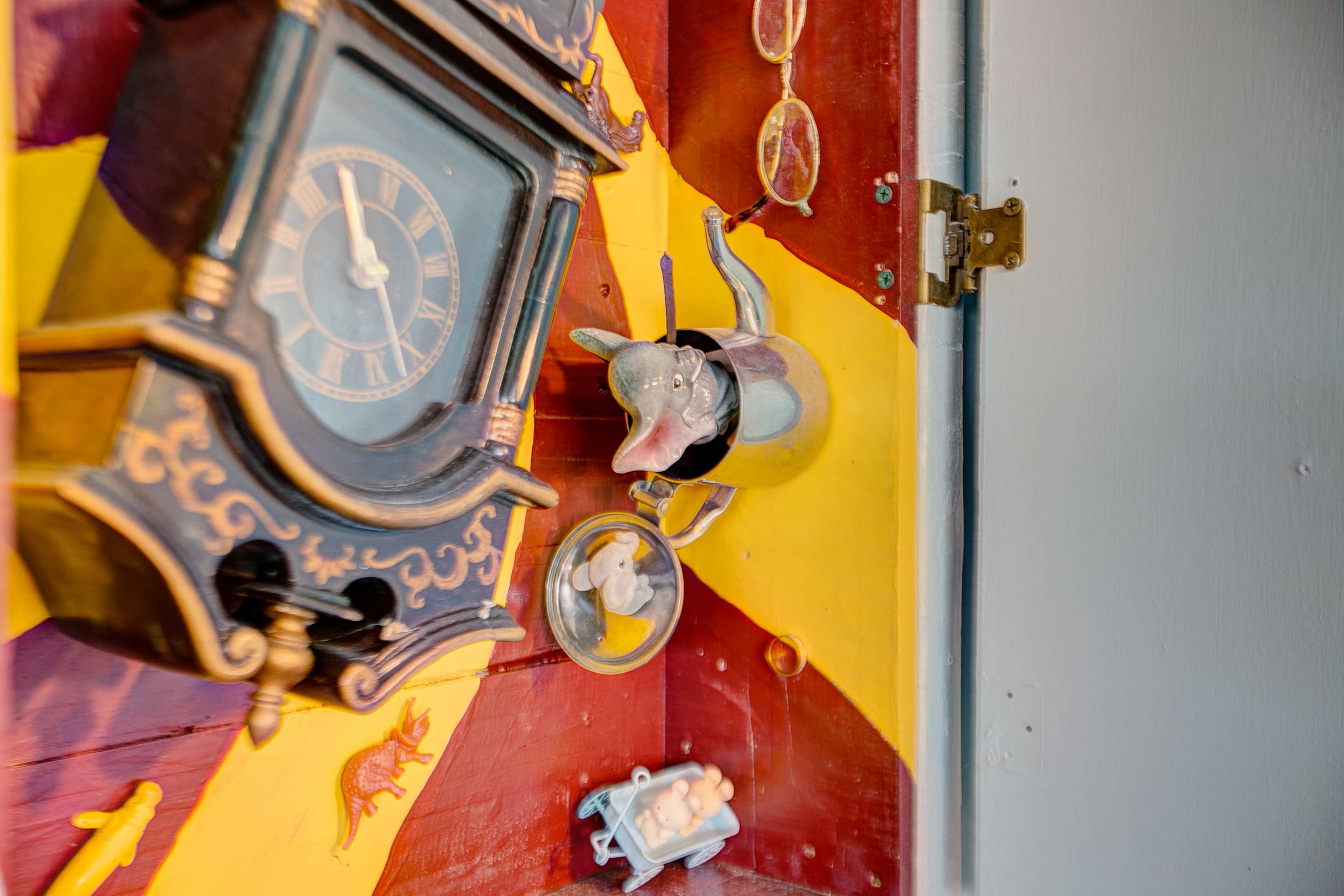 A colorful wall featuring a vintage clock, decorative glasses, a ceramic elephant, and small figurines on a red and yellow background.