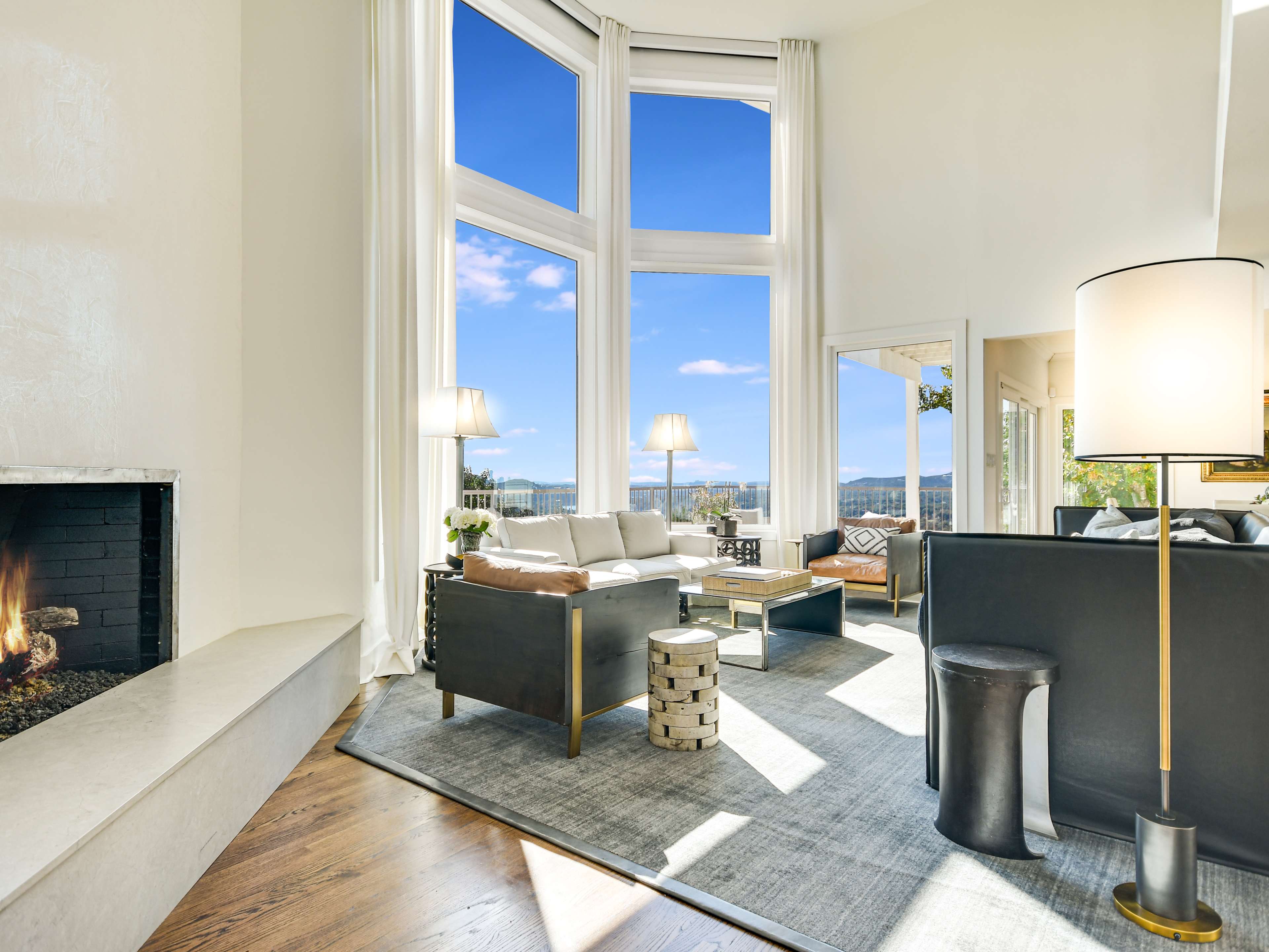A bright living room features large windows offering a view, a cozy fireplace, and modern furniture arranged around a central area rug.