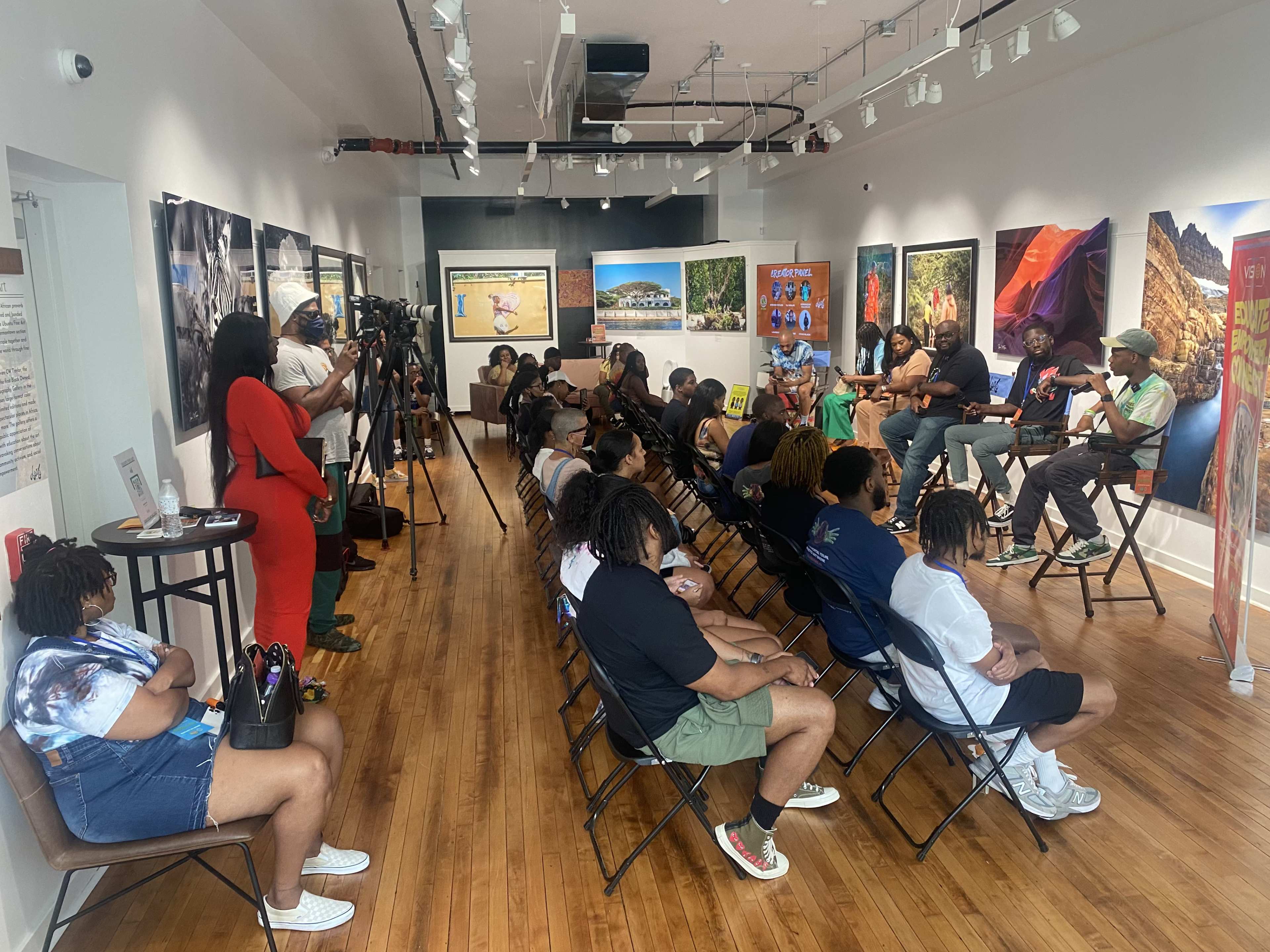 A group of people sits in rows of chairs in an art gallery, engaged in a discussion while cameras record the event.