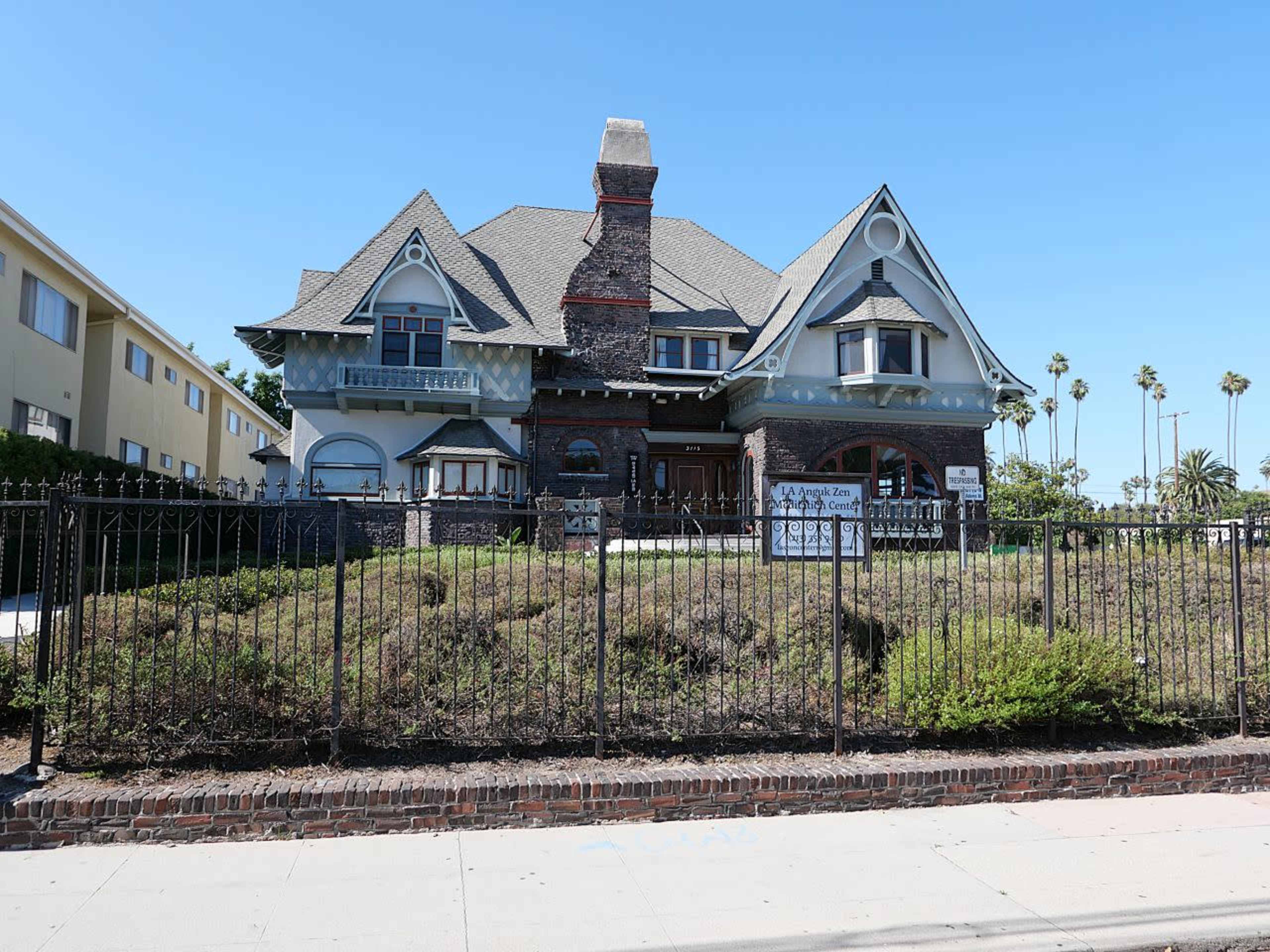The image shows a large, two-story Victorian-style house with a prominent chimney and ornate architectural details surrounded by a wrought-iron fence and overgrown landscaping.