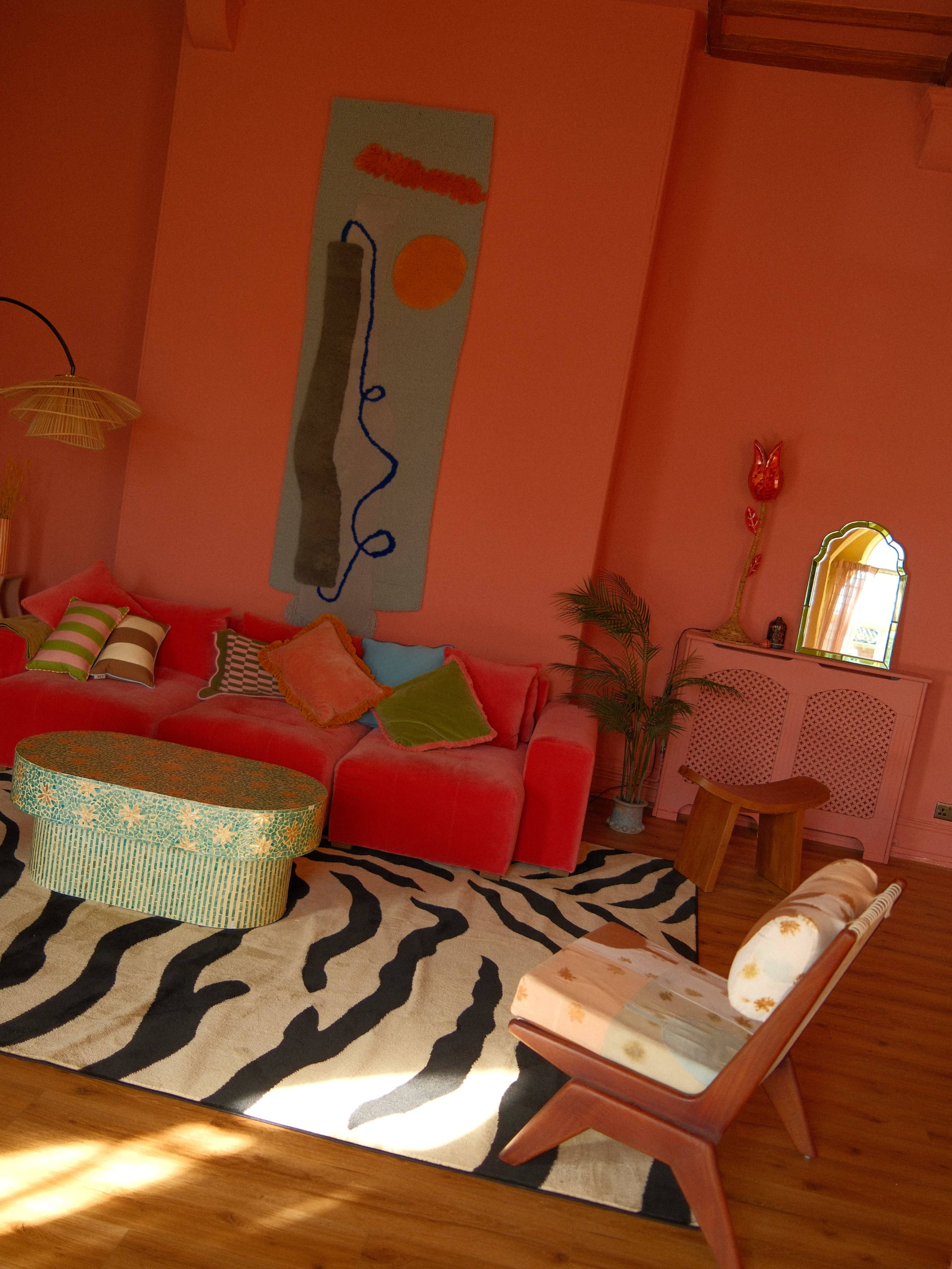 The image shows a brightly colored living room with a pink wall, a red sofa adorned with colorful pillows, a green patterned ottoman, and a zebra-striped rug on the wooden floor.