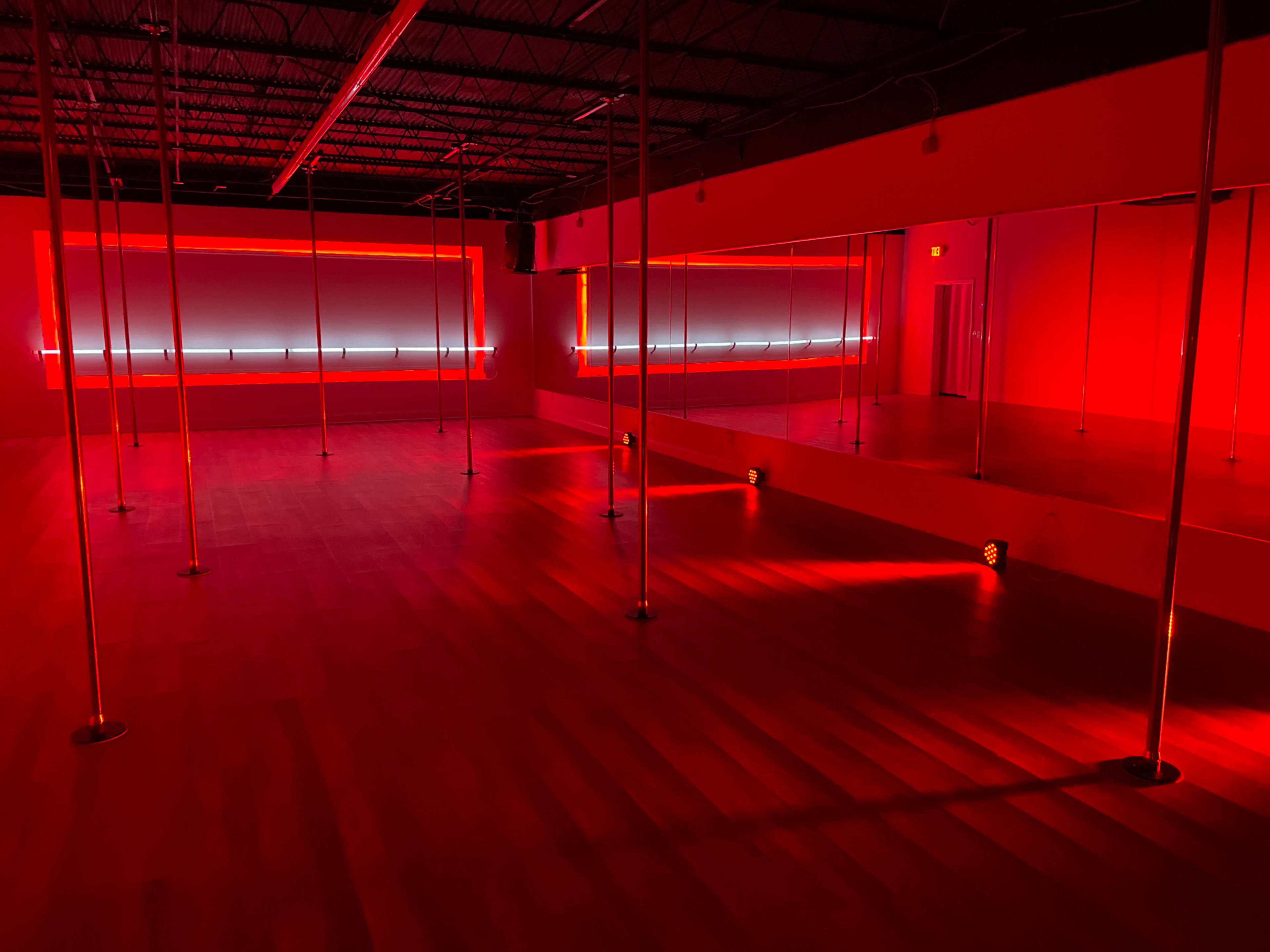 The image shows a spacious dance studio with polished wooden flooring and red-tinted lighting, featuring several vertical poles and mirrors.