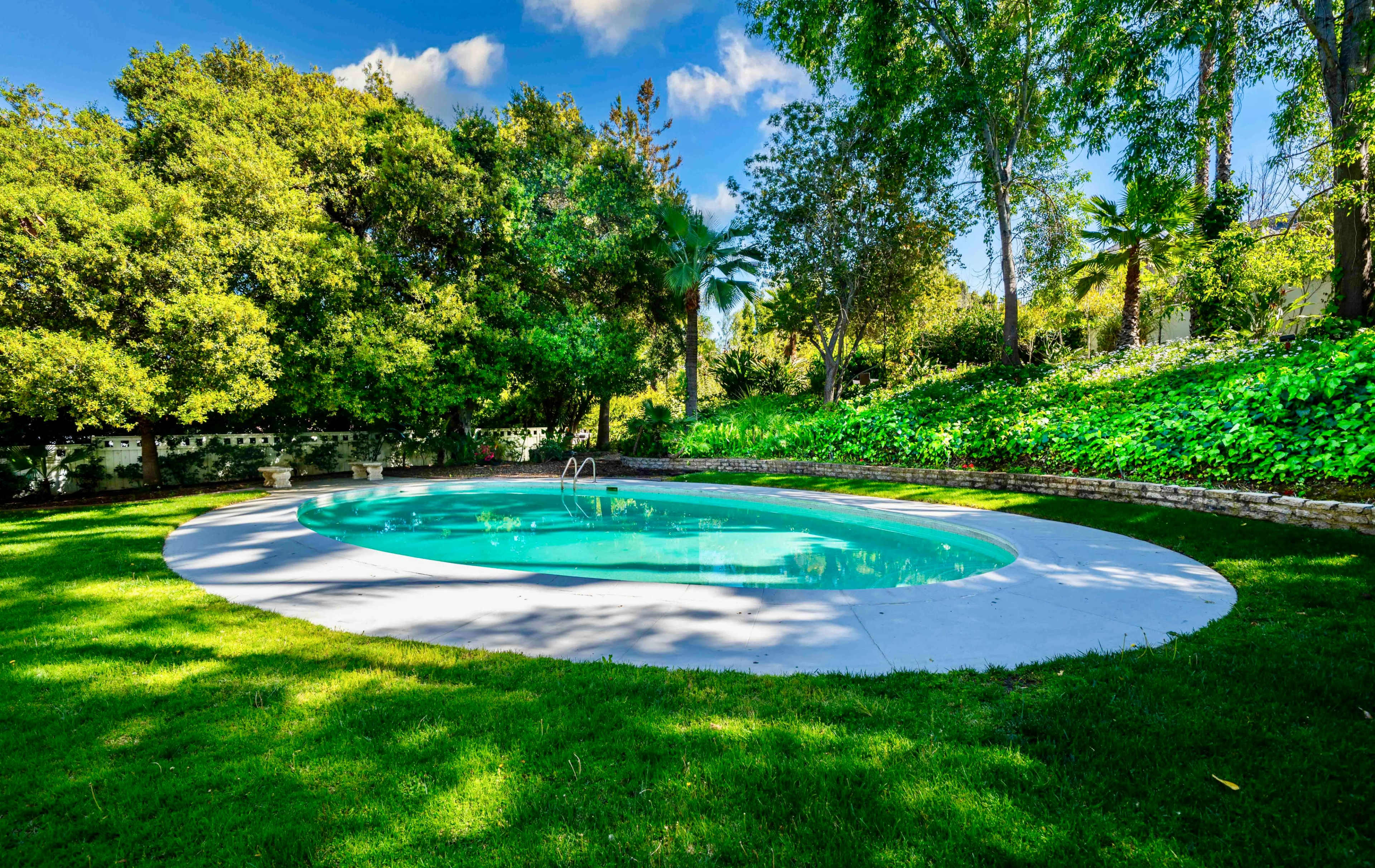 A curved swimming pool is surrounded by lush green grass and trees under a bright blue sky.