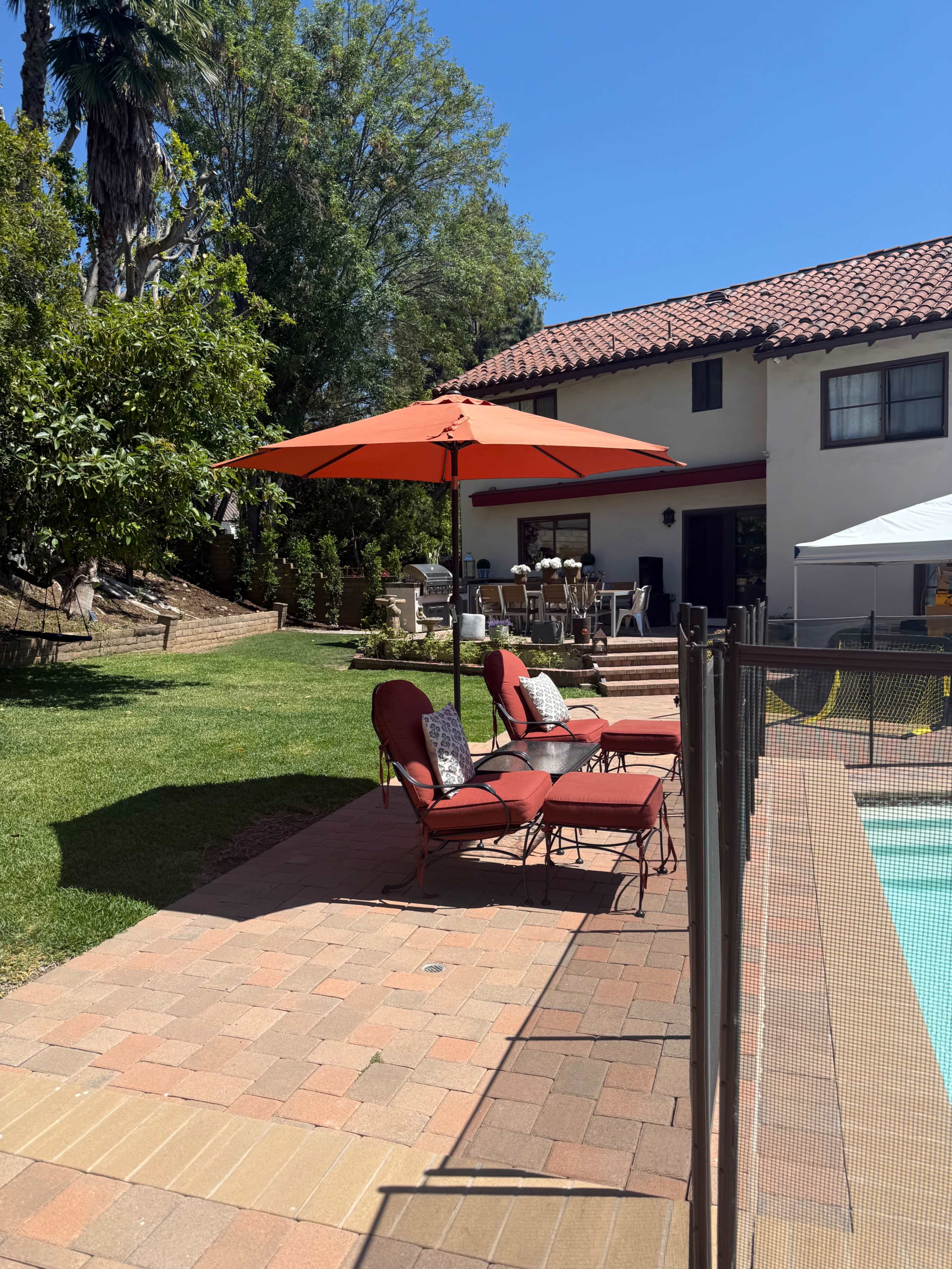 A patio area with red lounge chairs and an orange umbrella is situated near a swimming pool, with a house and outdoor dining area in the background.