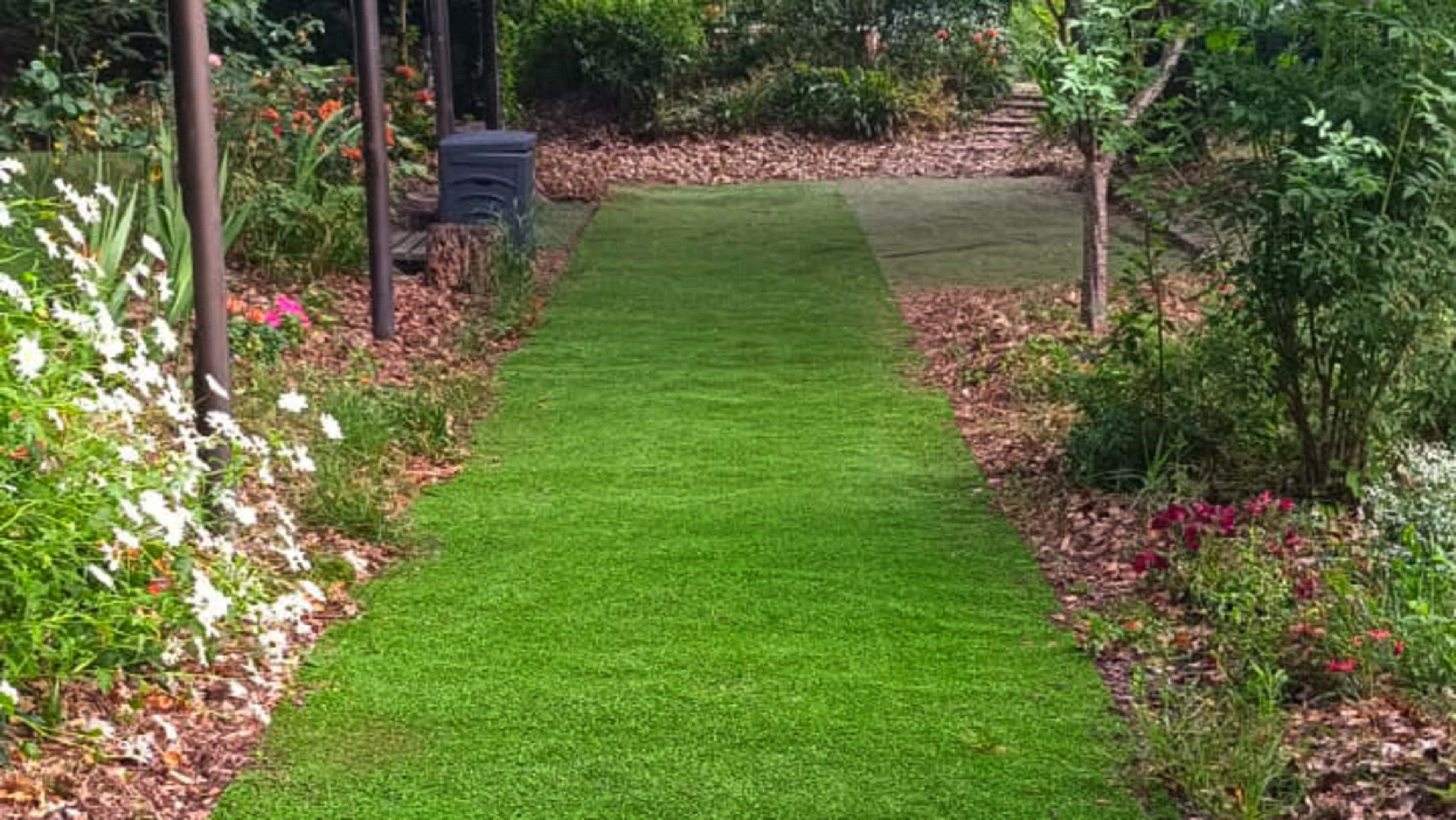 The image shows a green artificial turf pathway bordered by flower beds and trees in a garden setting.