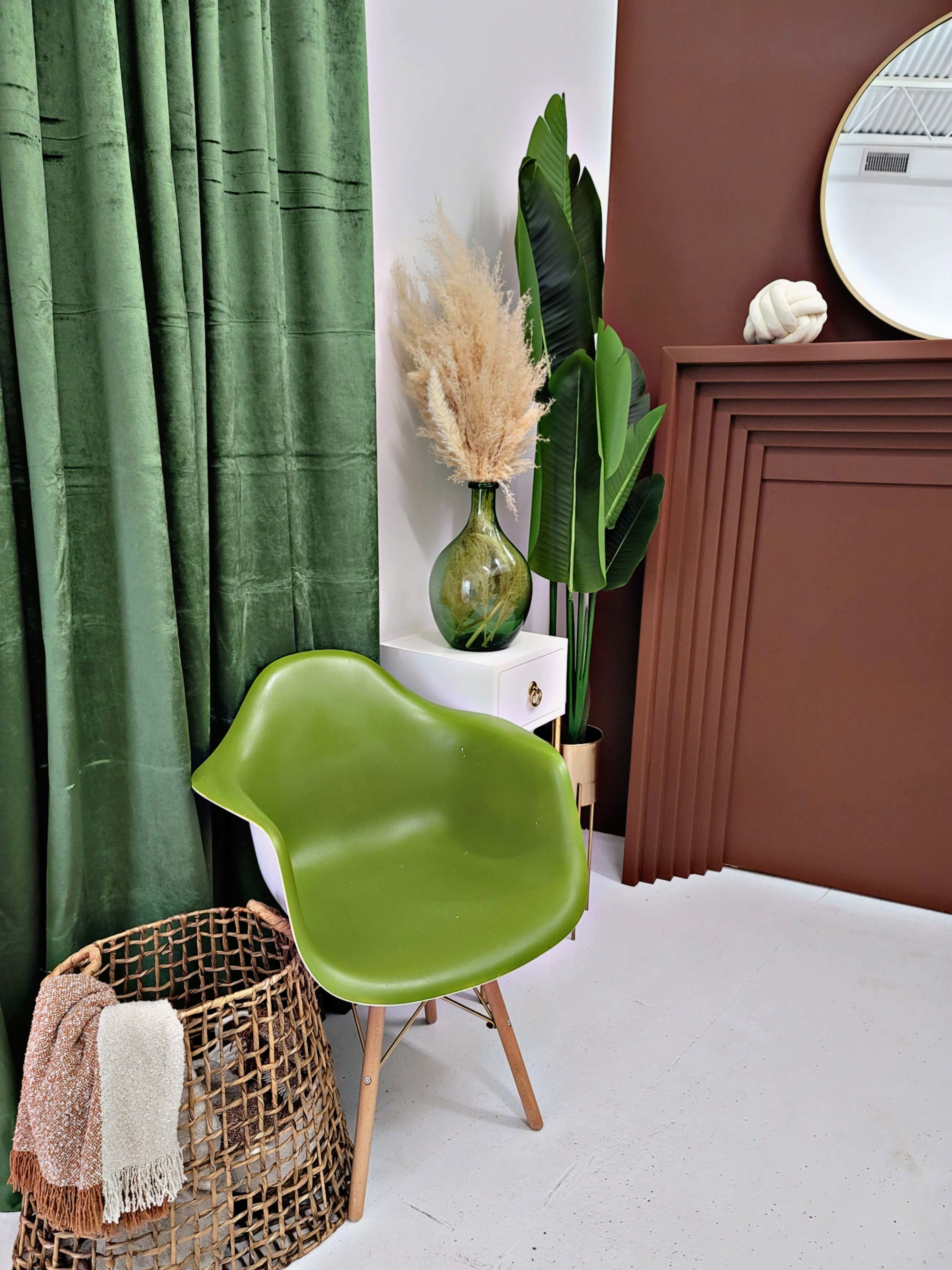 The image features a green chair beside a woven basket, with green curtains, a decorative vase containing dried plants, and a brown accent wall.