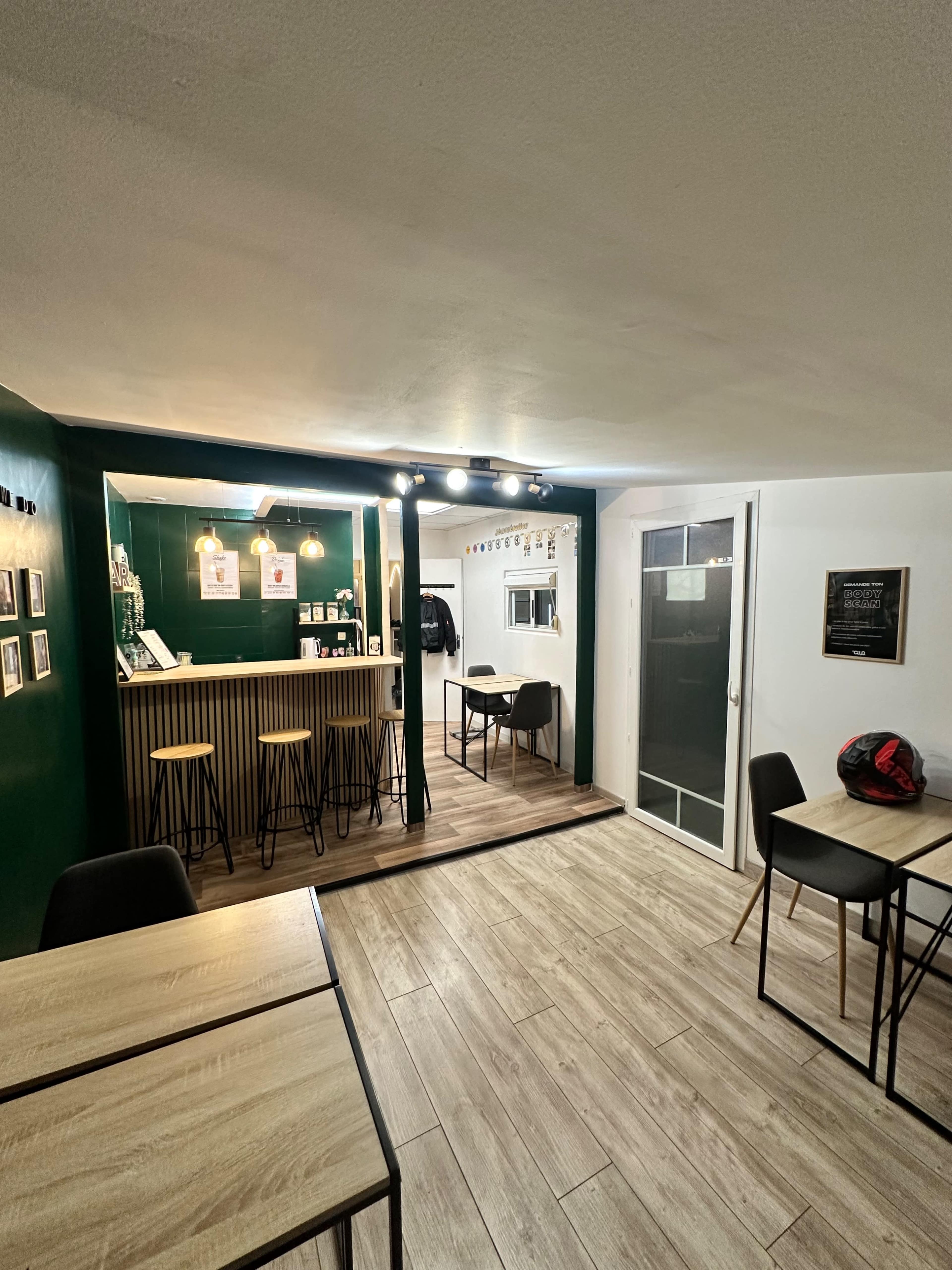 The image shows a cozy café interior with wooden tables, black chairs, and a small service counter illuminated with warm lights.