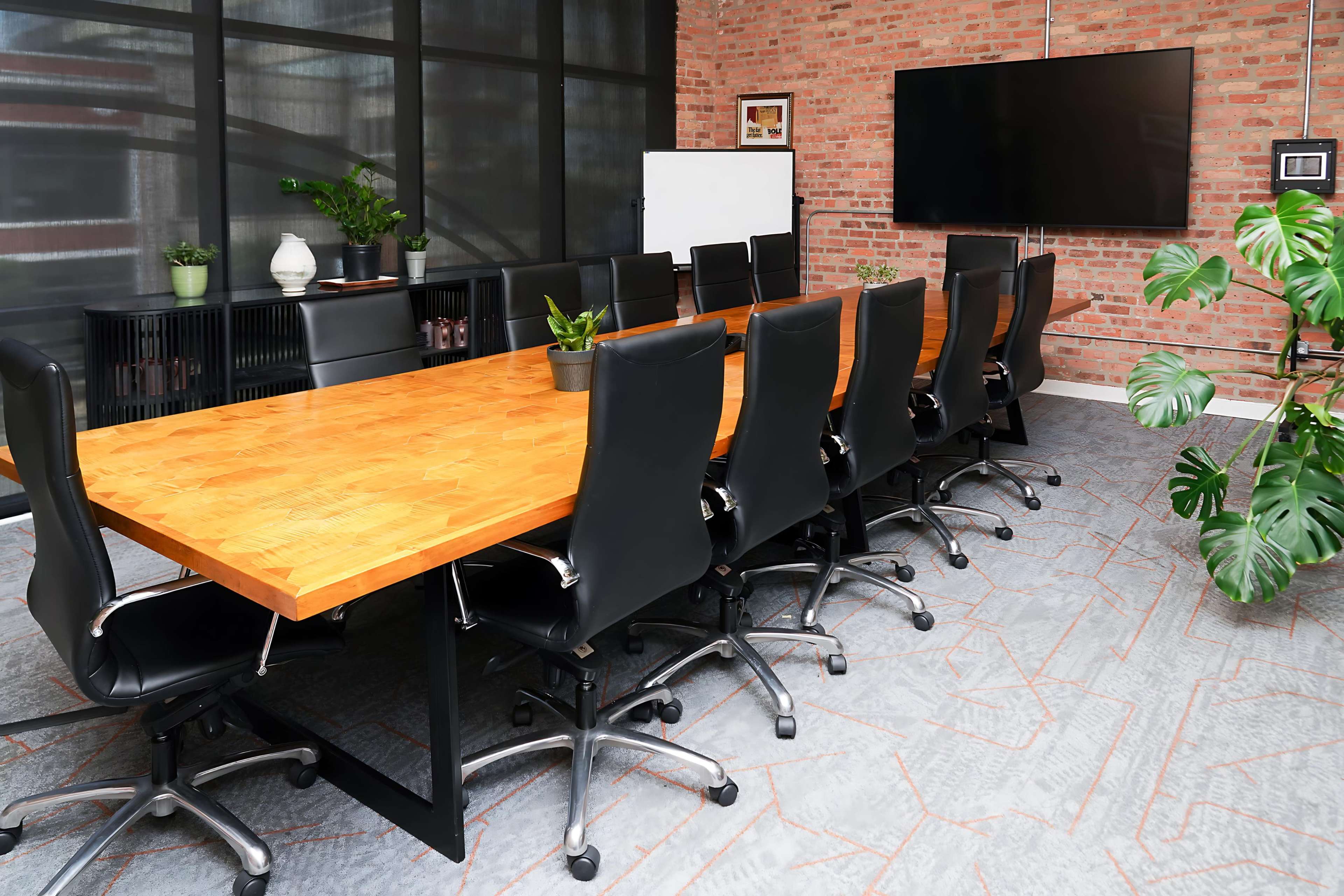The image shows a modern conference room with a long wooden table surrounded by black office chairs, large windows, a television screen, and green plants.