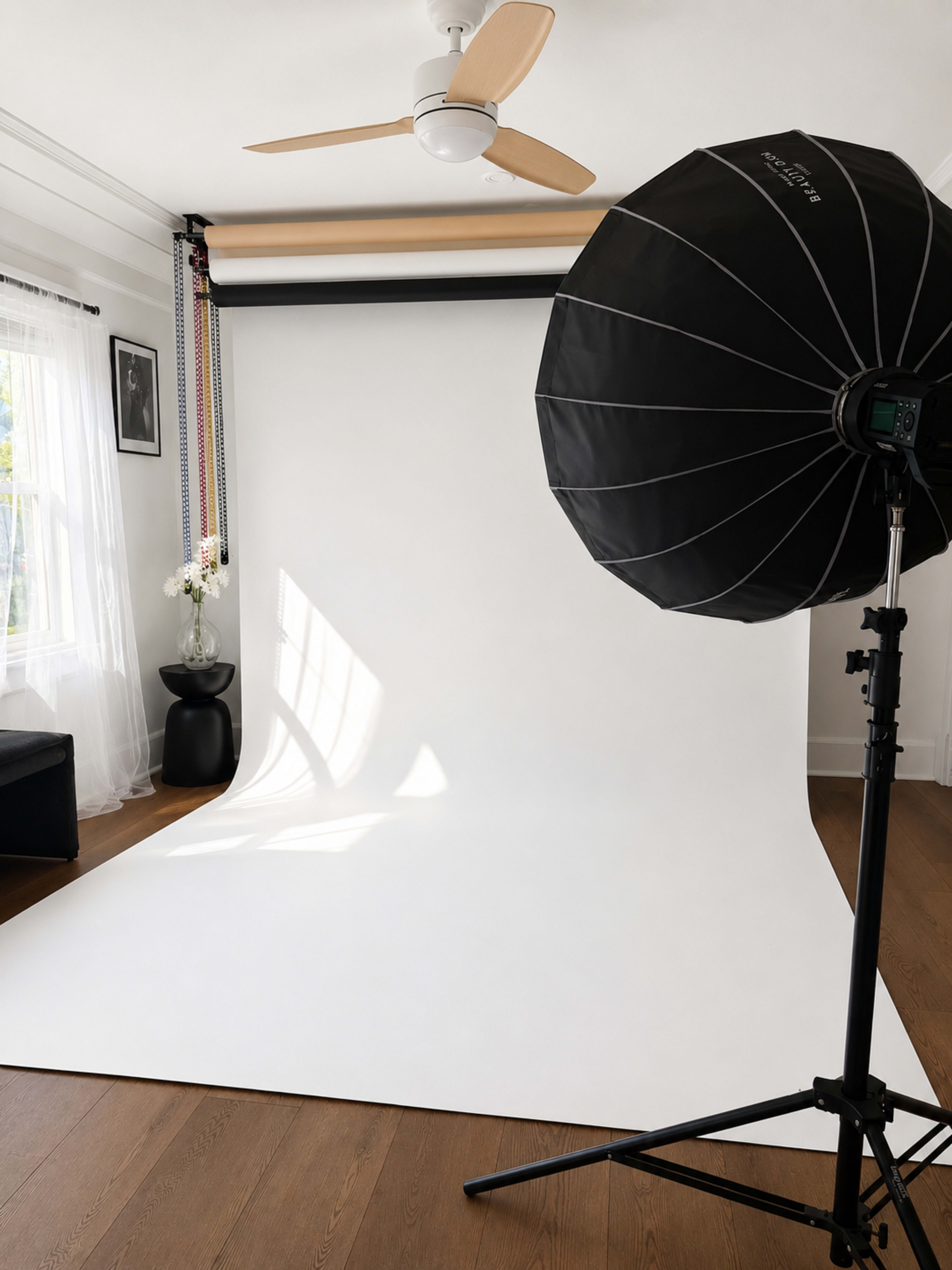 The image shows a photography studio with a large softbox light and a white backdrop in an uncluttered room featuring a ceiling fan and a small plant.