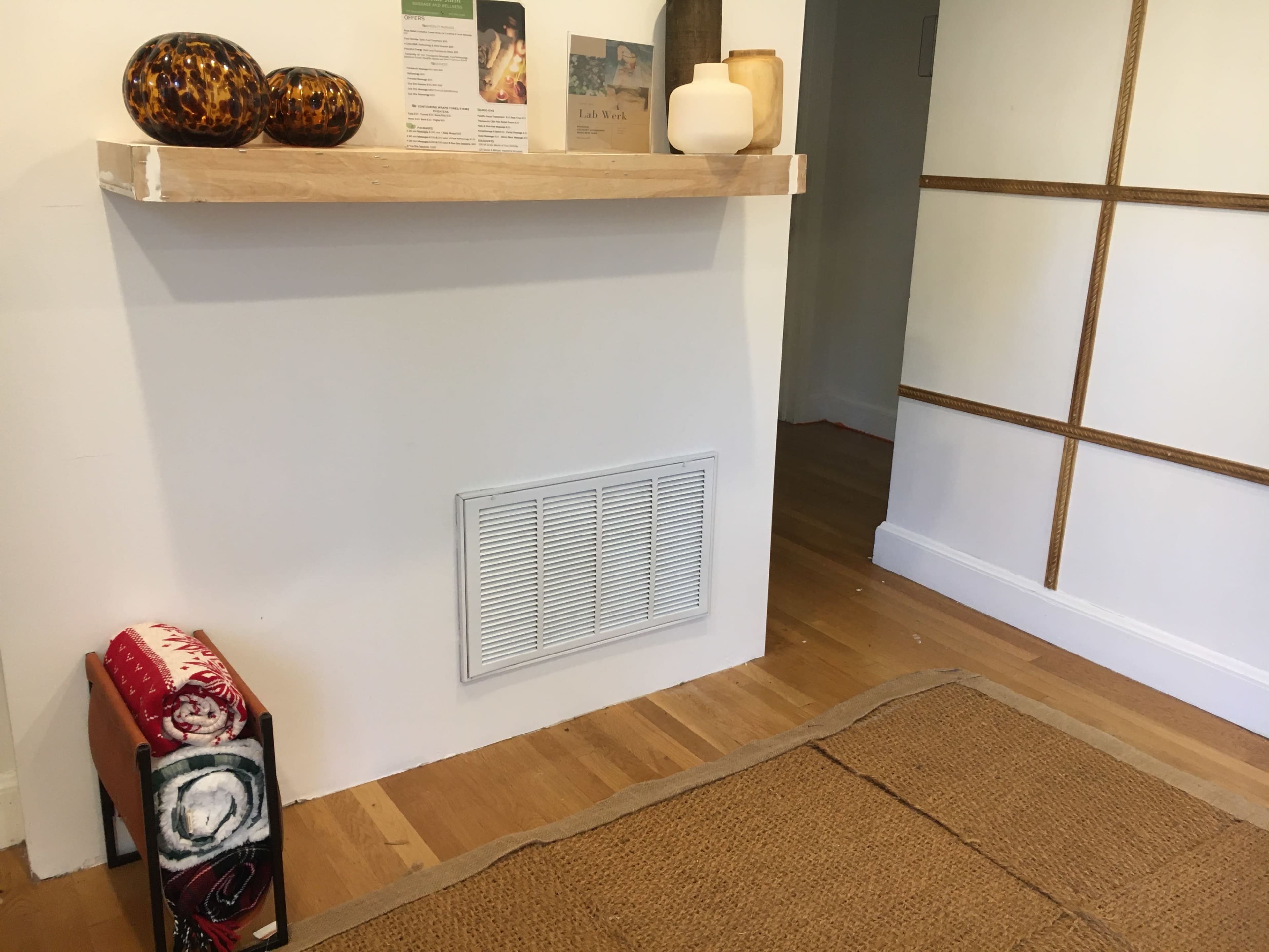 A wooden shelf with two decorative items is mounted on a white wall above a ventilation grate, adjacent to a woven rug on the floor.