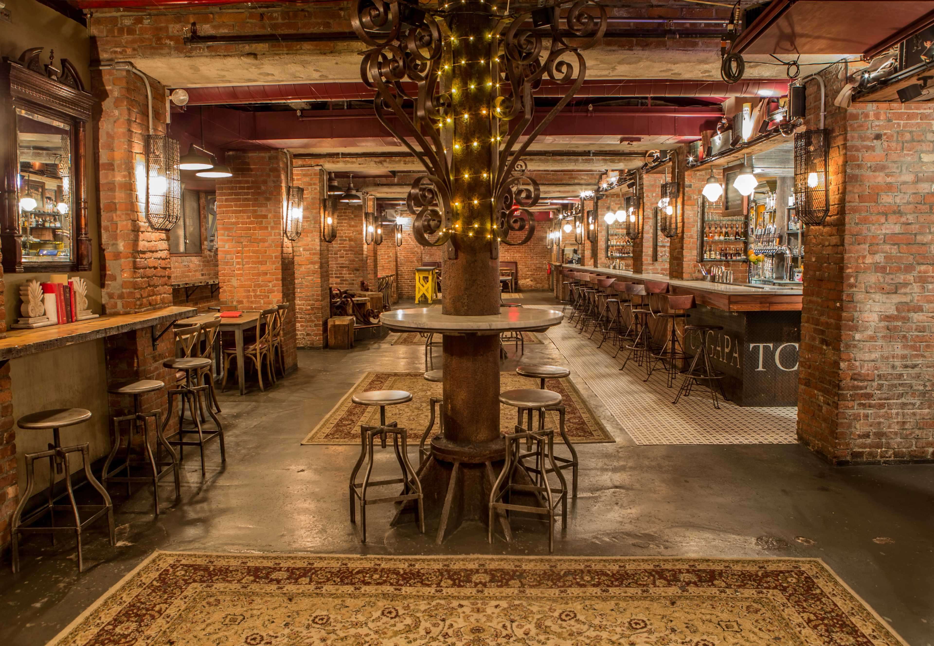 The image shows a rustic bar interior with exposed brick walls, wooden seating, and a central column decorated with string lights.