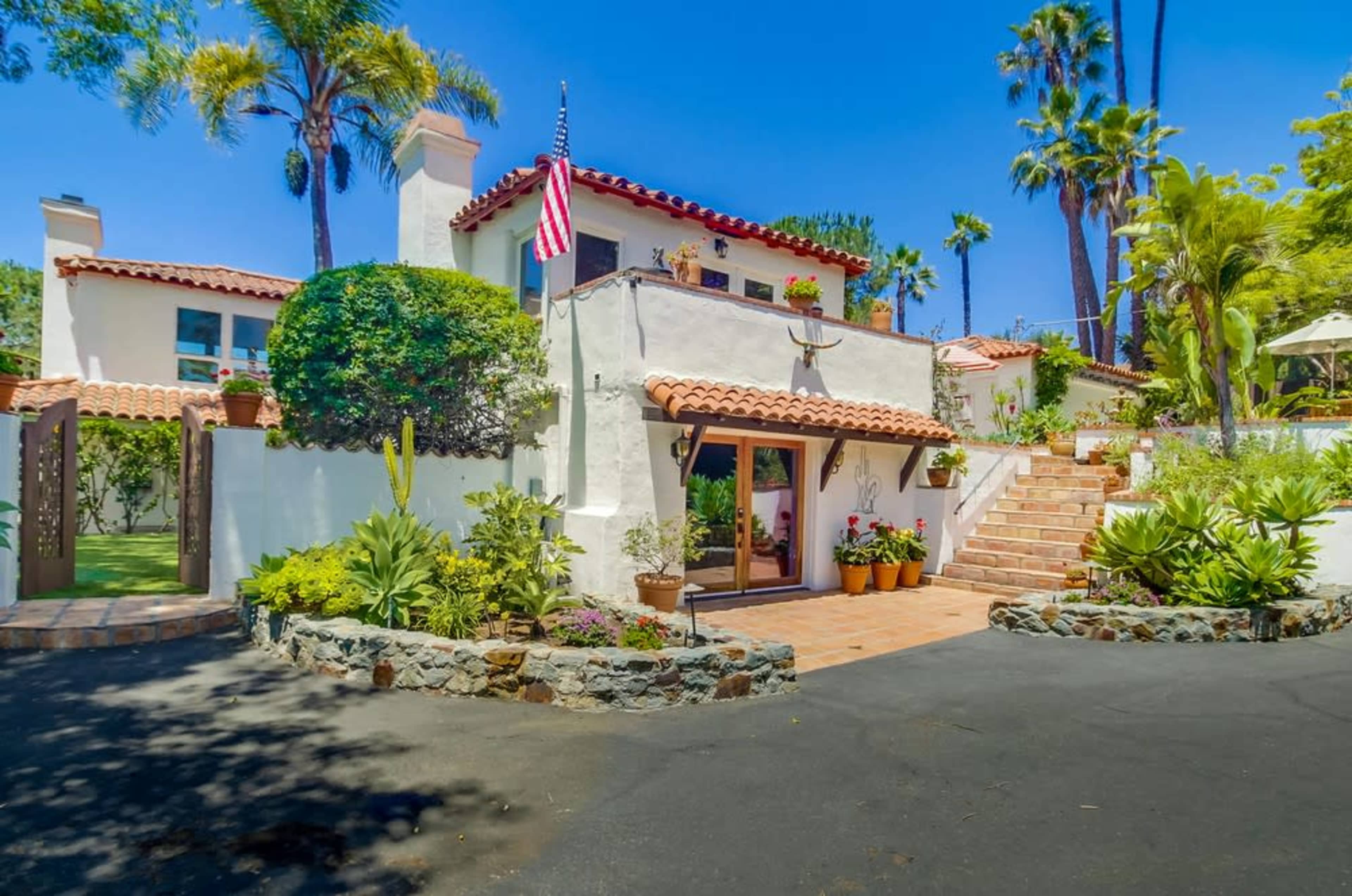 The image shows a Mediterranean-style house with a red-tiled roof, surrounded by various plants and palm trees, along with a brick pathway leading to the entrance.