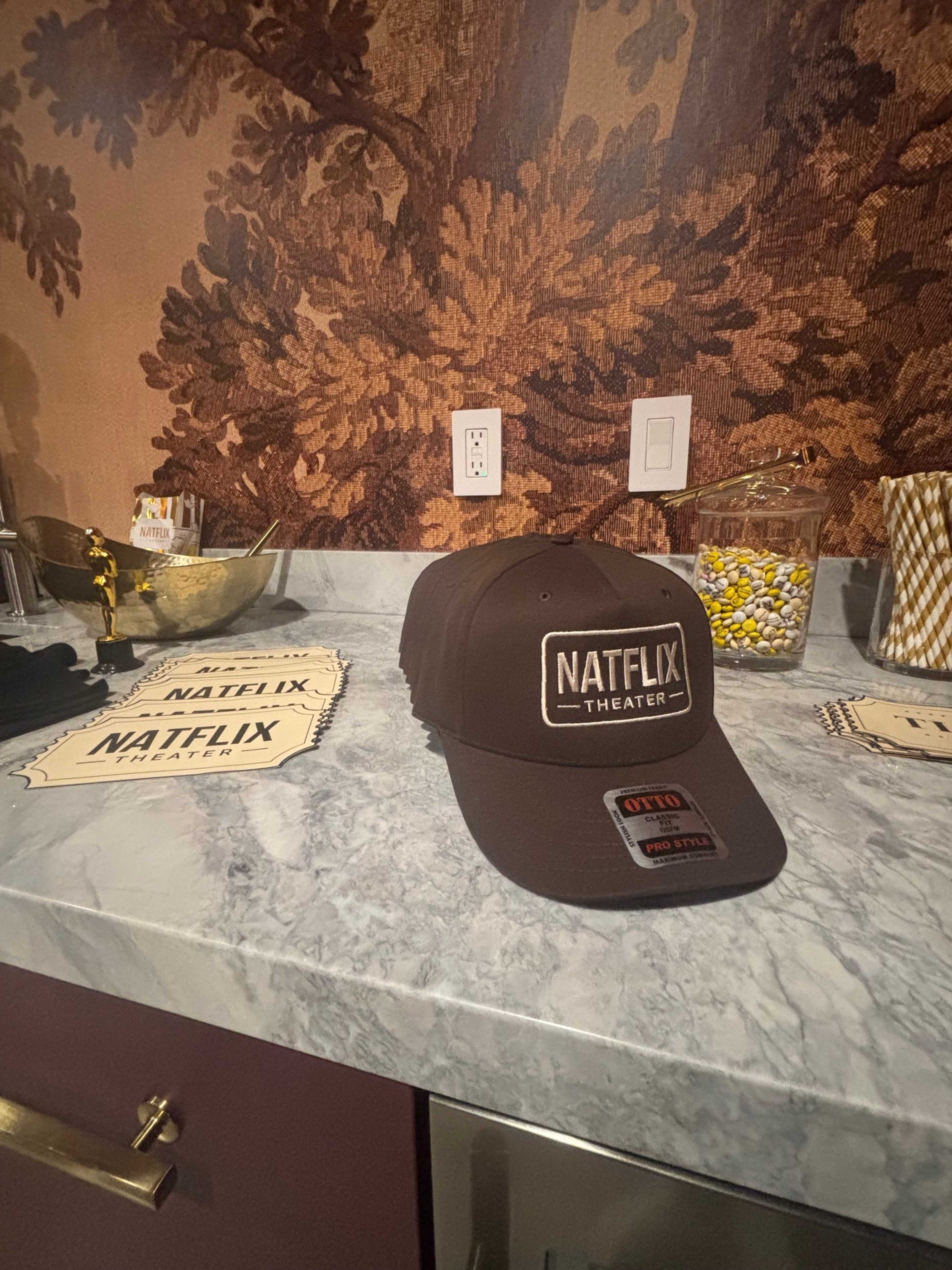 The image shows a brown cap with the text "NATFLIX THEATER" resting on a marble countertop beside decorative items and a bowl of colorful candy.