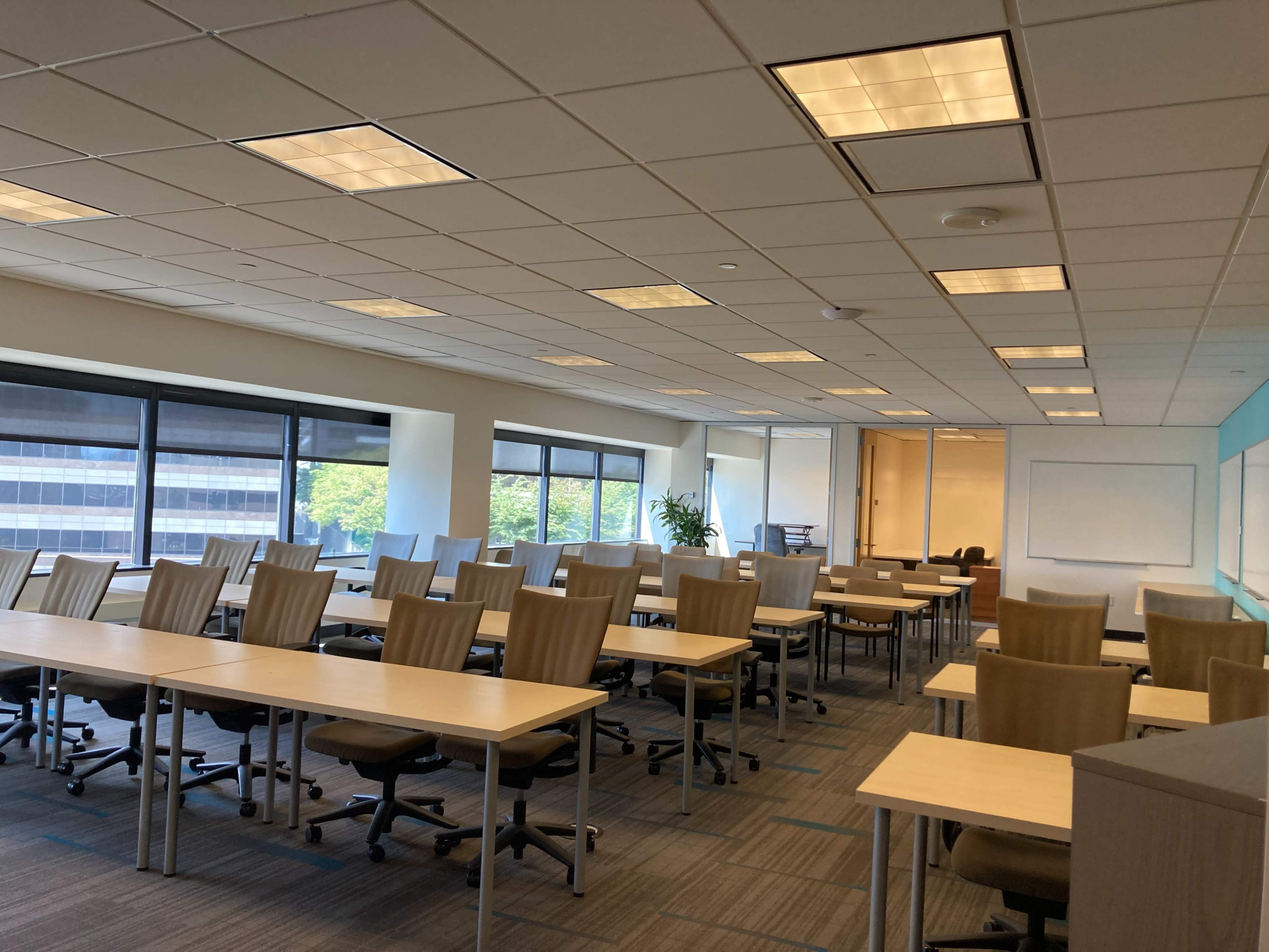 A spacious, modern classroom with multiple rows of desks and chairs, large windows, and a whiteboard.