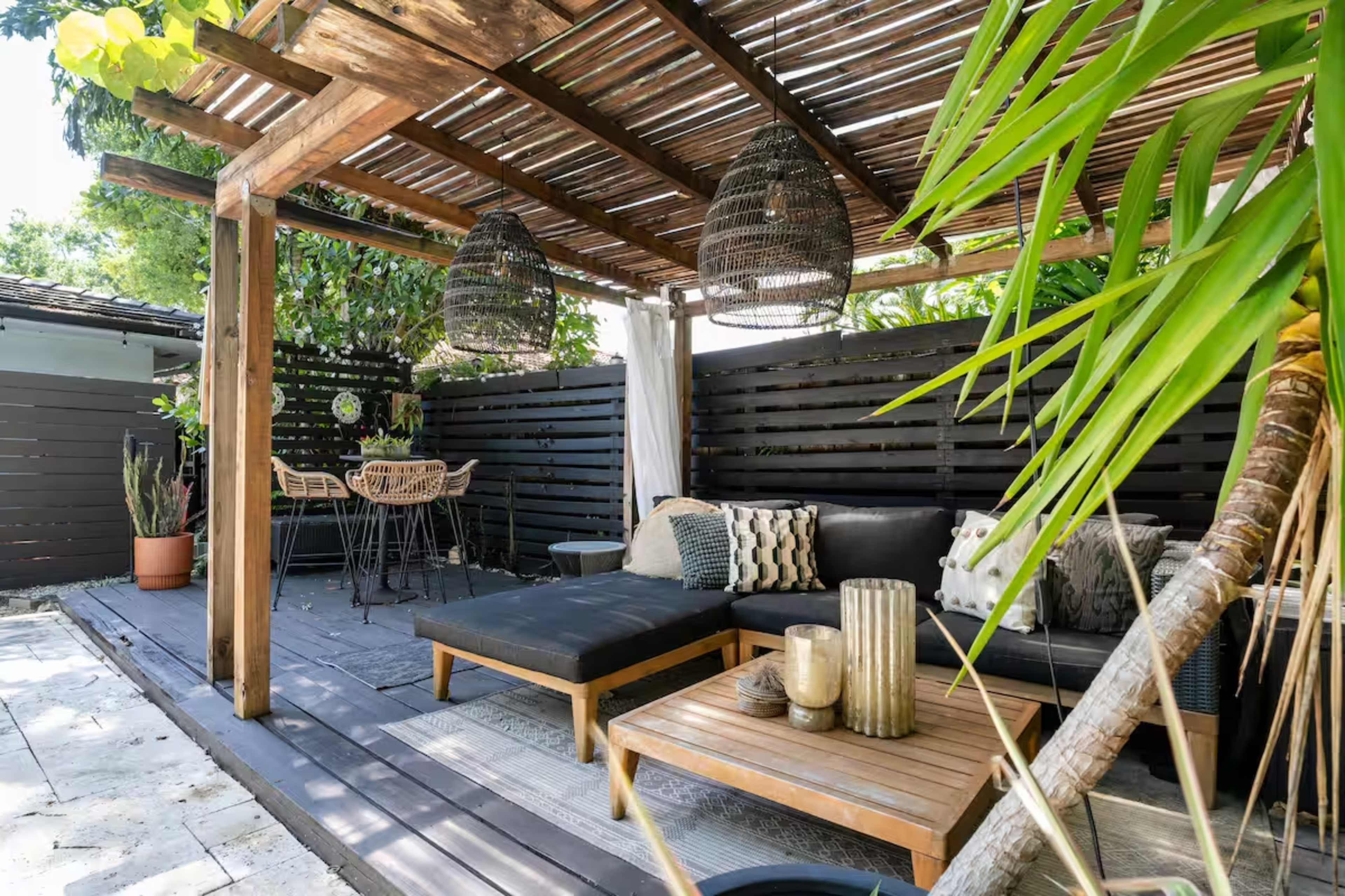The image shows a modern outdoor seating area with a wooden pergola, featuring a sectional sofa, a coffee table, and two hanging light fixtures.