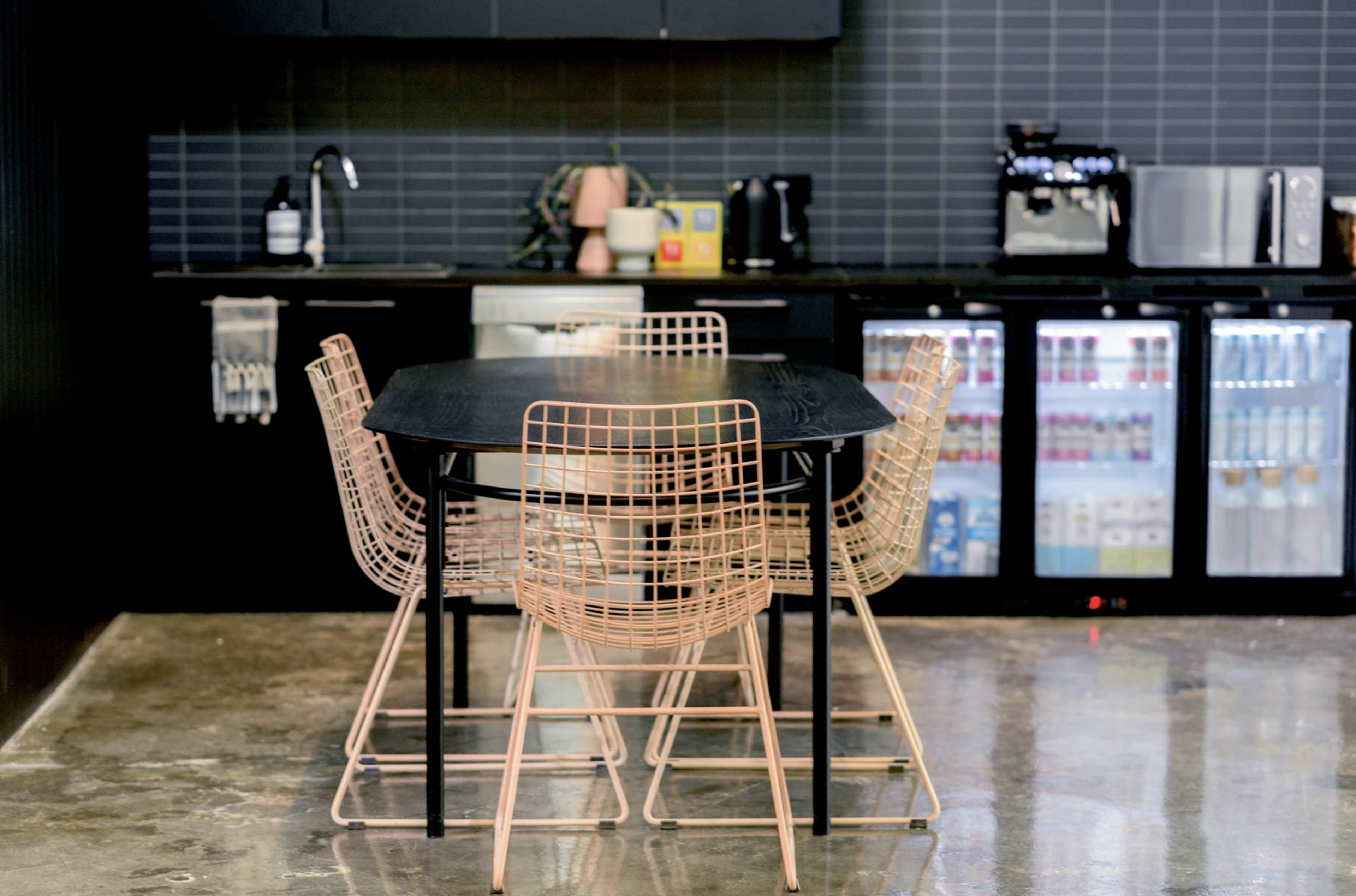 A black table with four wireframe chairs is set in a modern kitchen with dark cabinetry and stainless steel appliances.