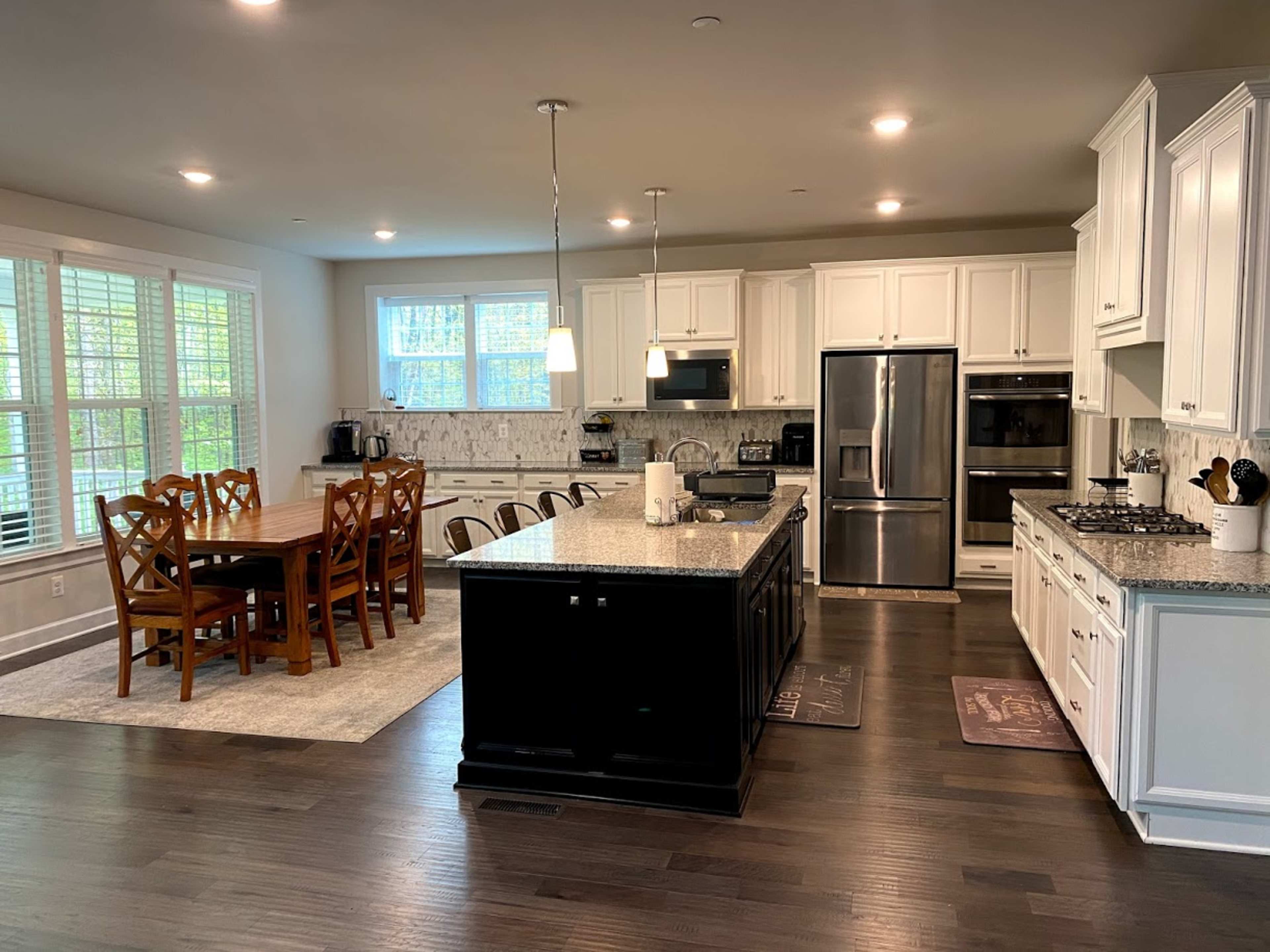 A modern kitchen with white cabinets, a large island with dark granite countertops, a dining table with six wooden chairs, and stainless steel appliances.