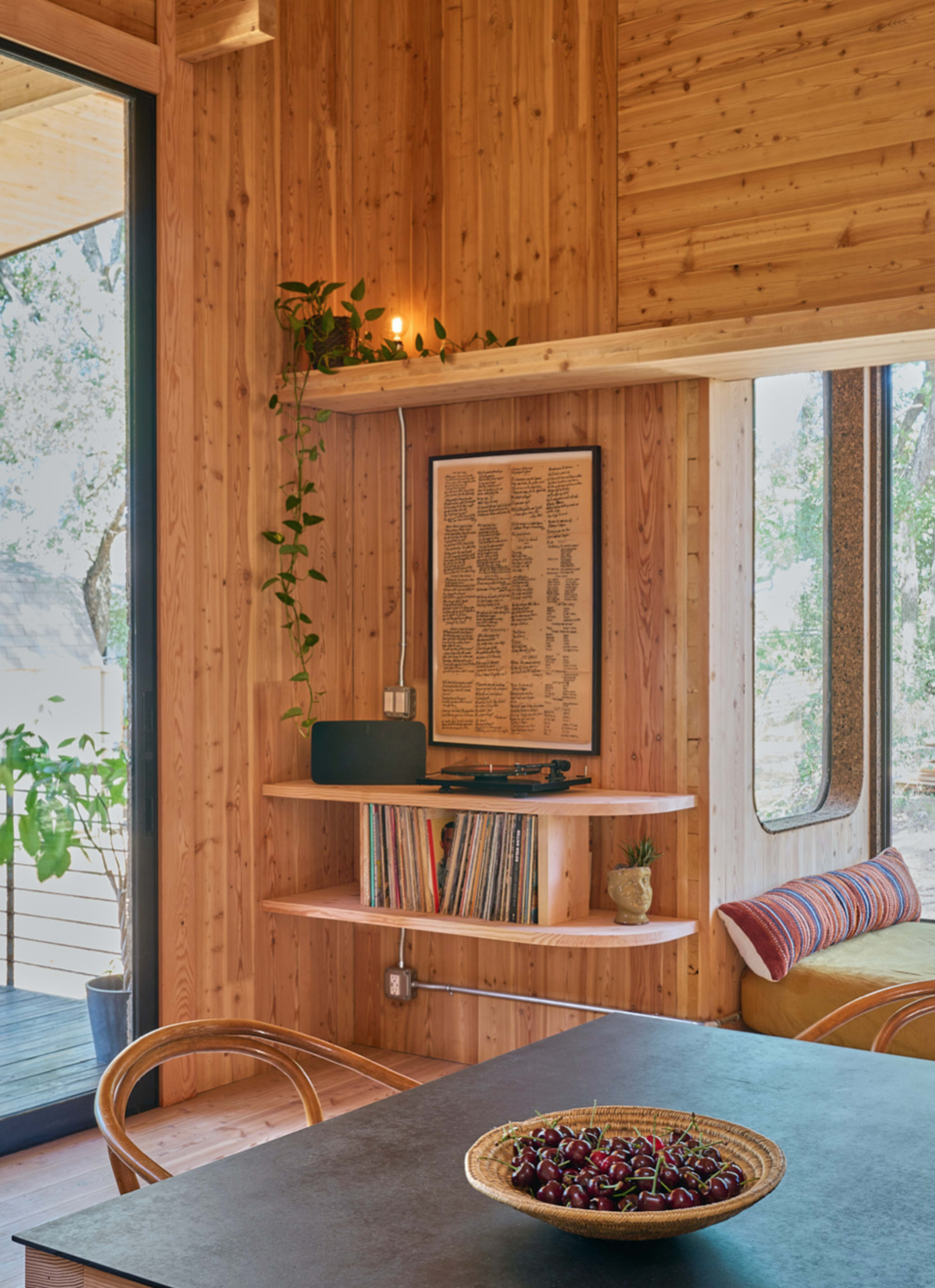 A wooden interior featuring shelves with books and plants, a record player, and a round bowl of cherries on a dark table.