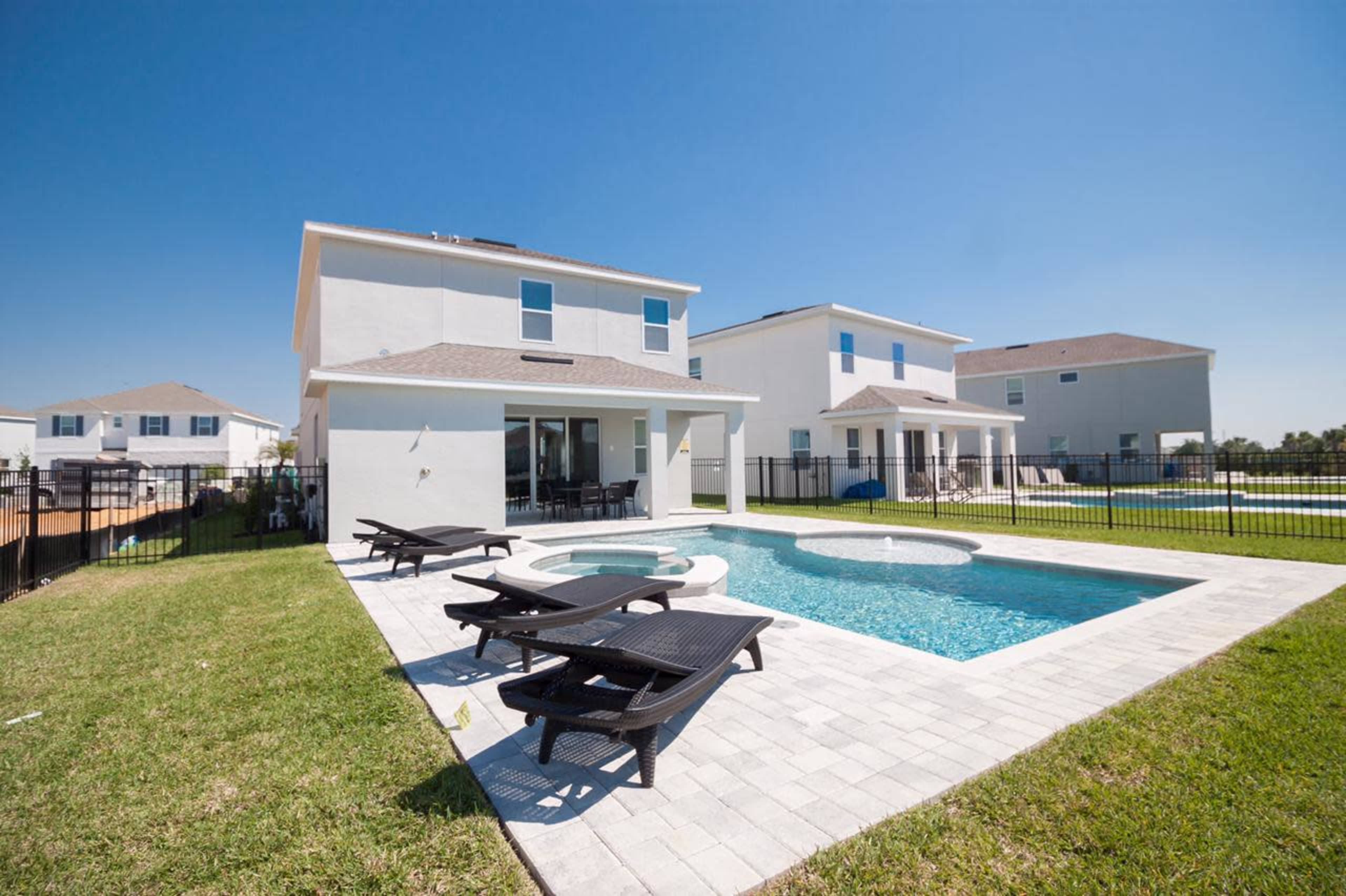 A backyard with a swimming pool surrounded by lounge chairs, two houses, and a fenced area under a clear blue sky.