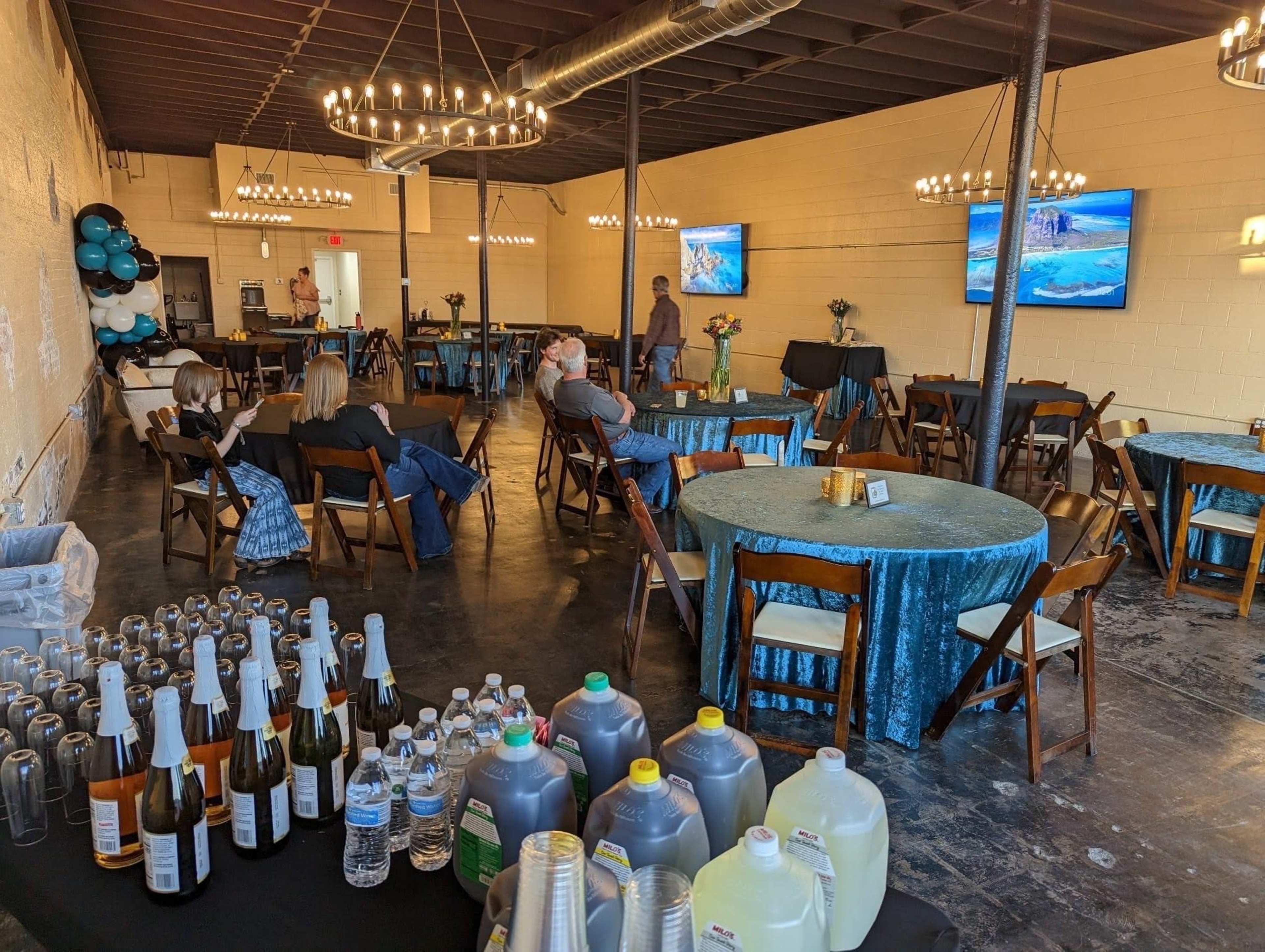 A spacious event venue features round tables with teal tablecloths, seated guests, and refreshments displayed in the foreground.
