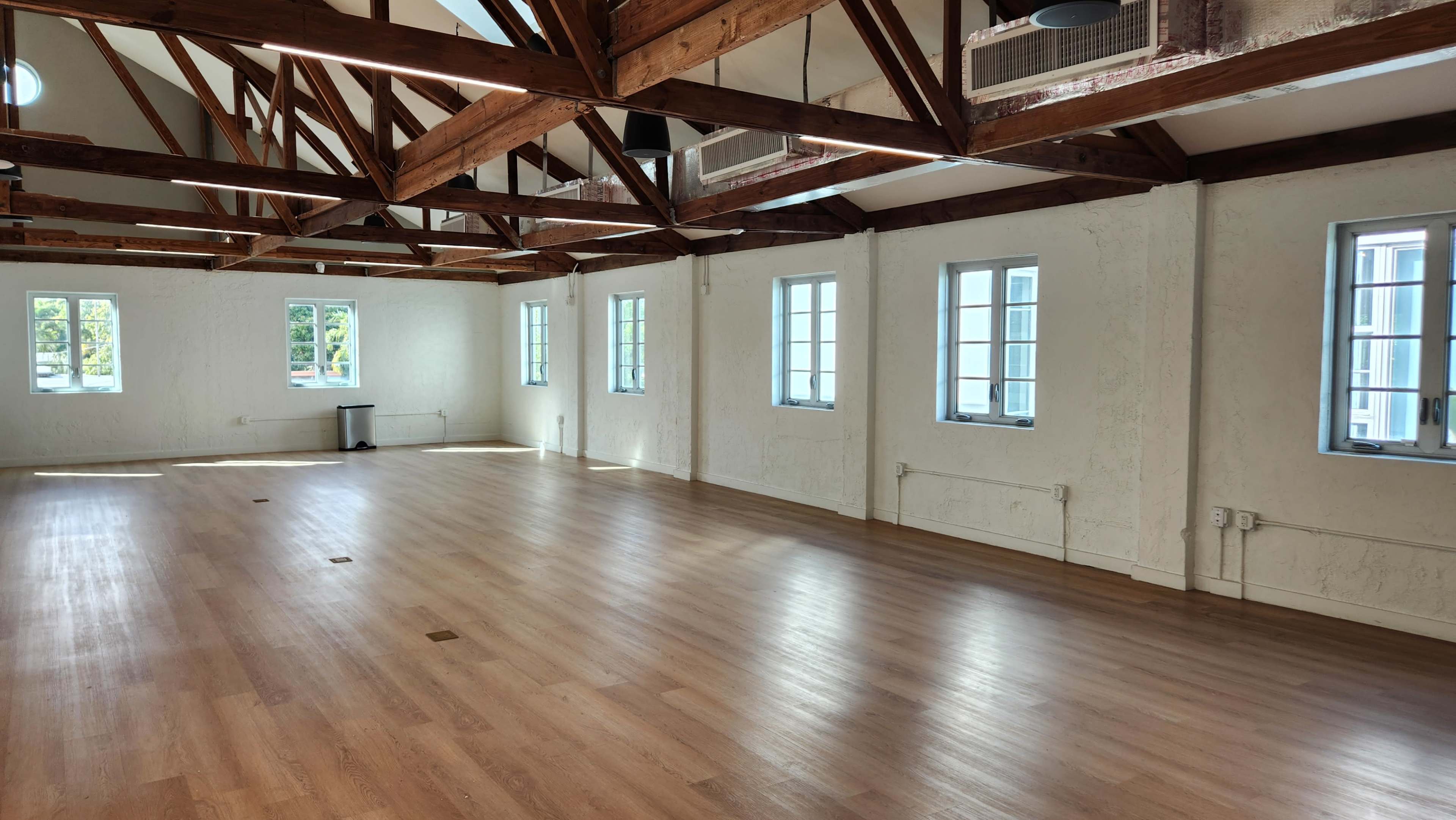 The image shows a spacious, well-lit interior with wooden beams and multiple windows, featuring a hardwood floor and minimal furnishings.