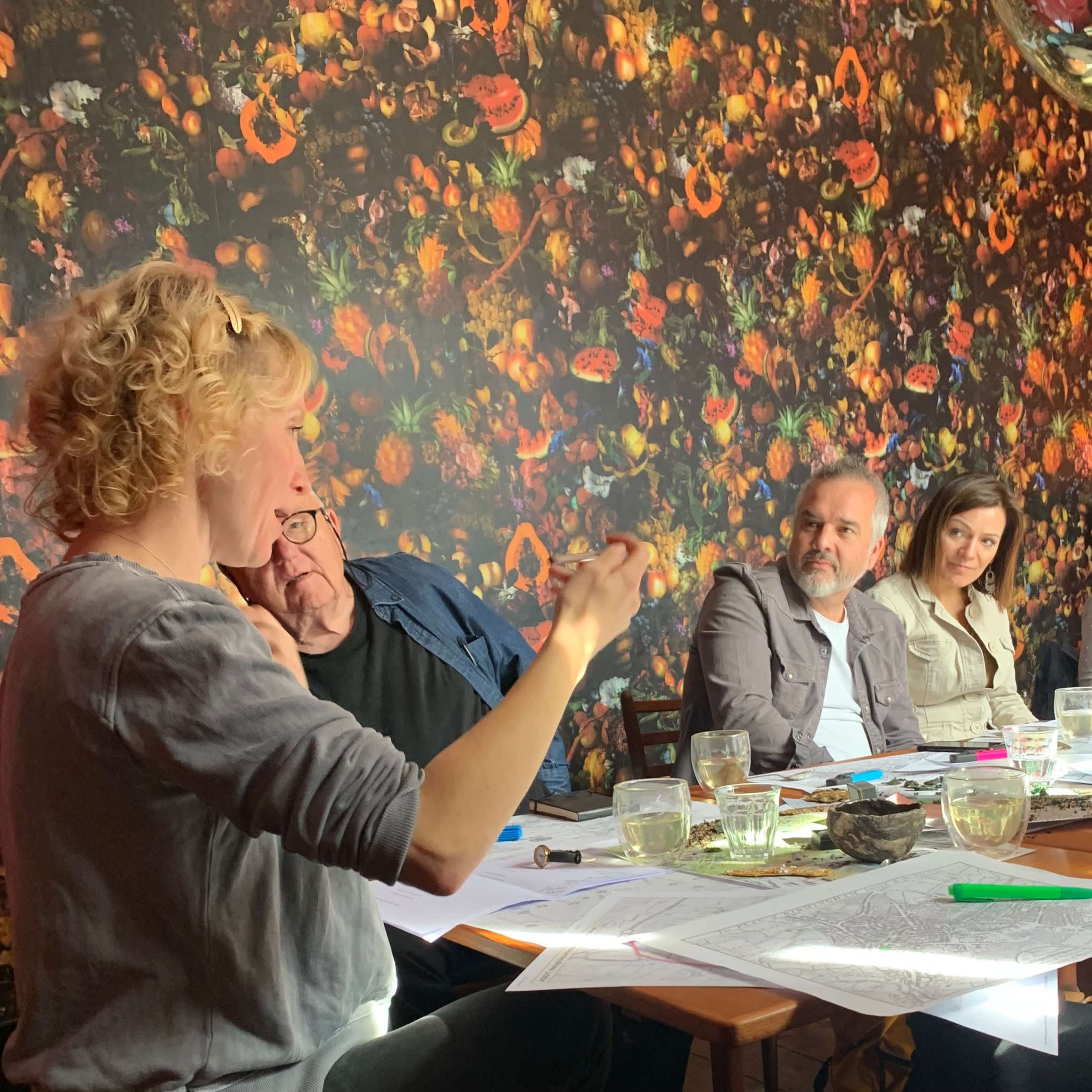 A group of four people is seated around a table, discussing while surrounded by a flower-covered wall.