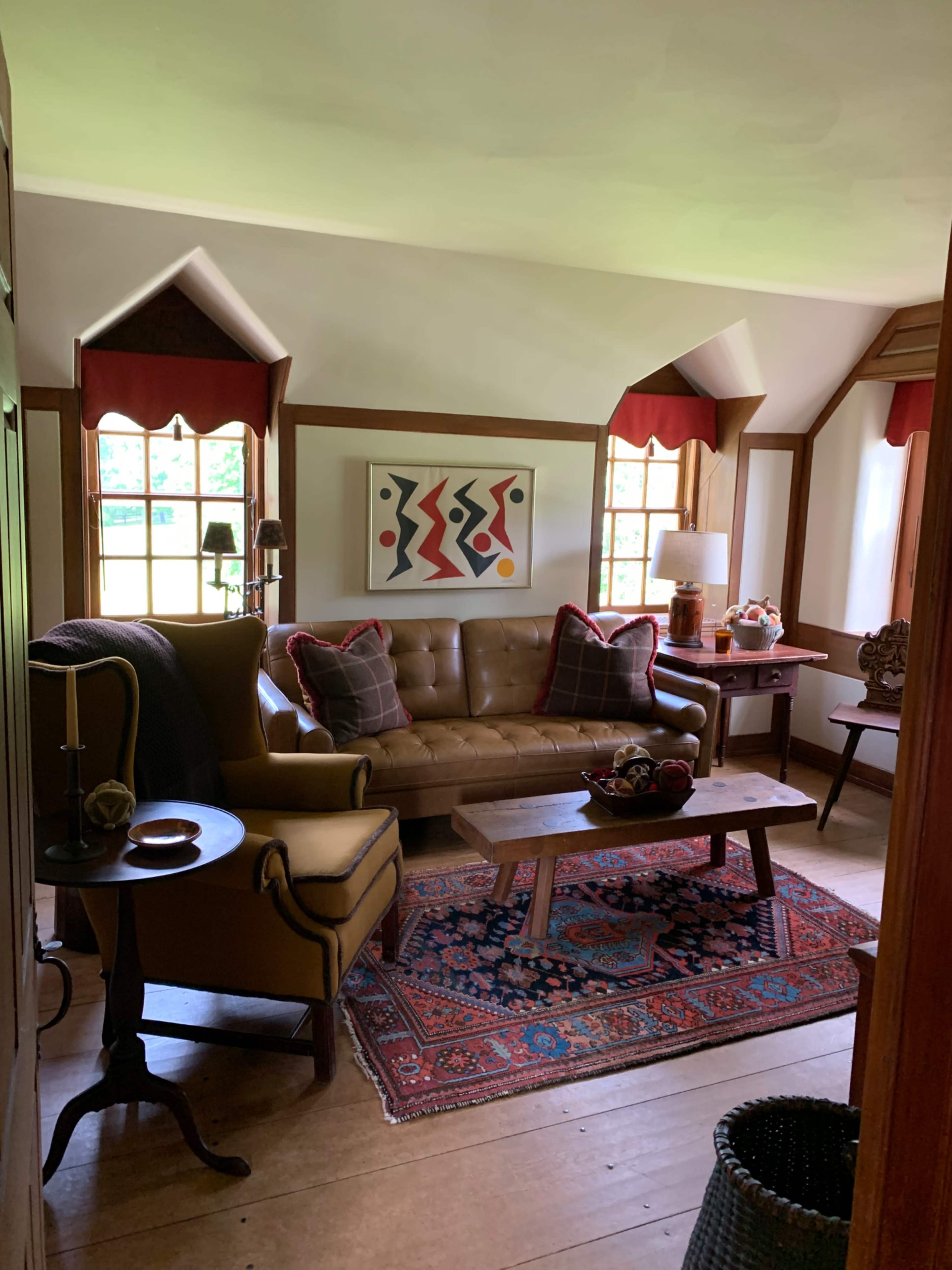 A cozy living room features a brown leather sofa, a wooden coffee table on an area rug, and colorful wall art, with light streaming through window panes.