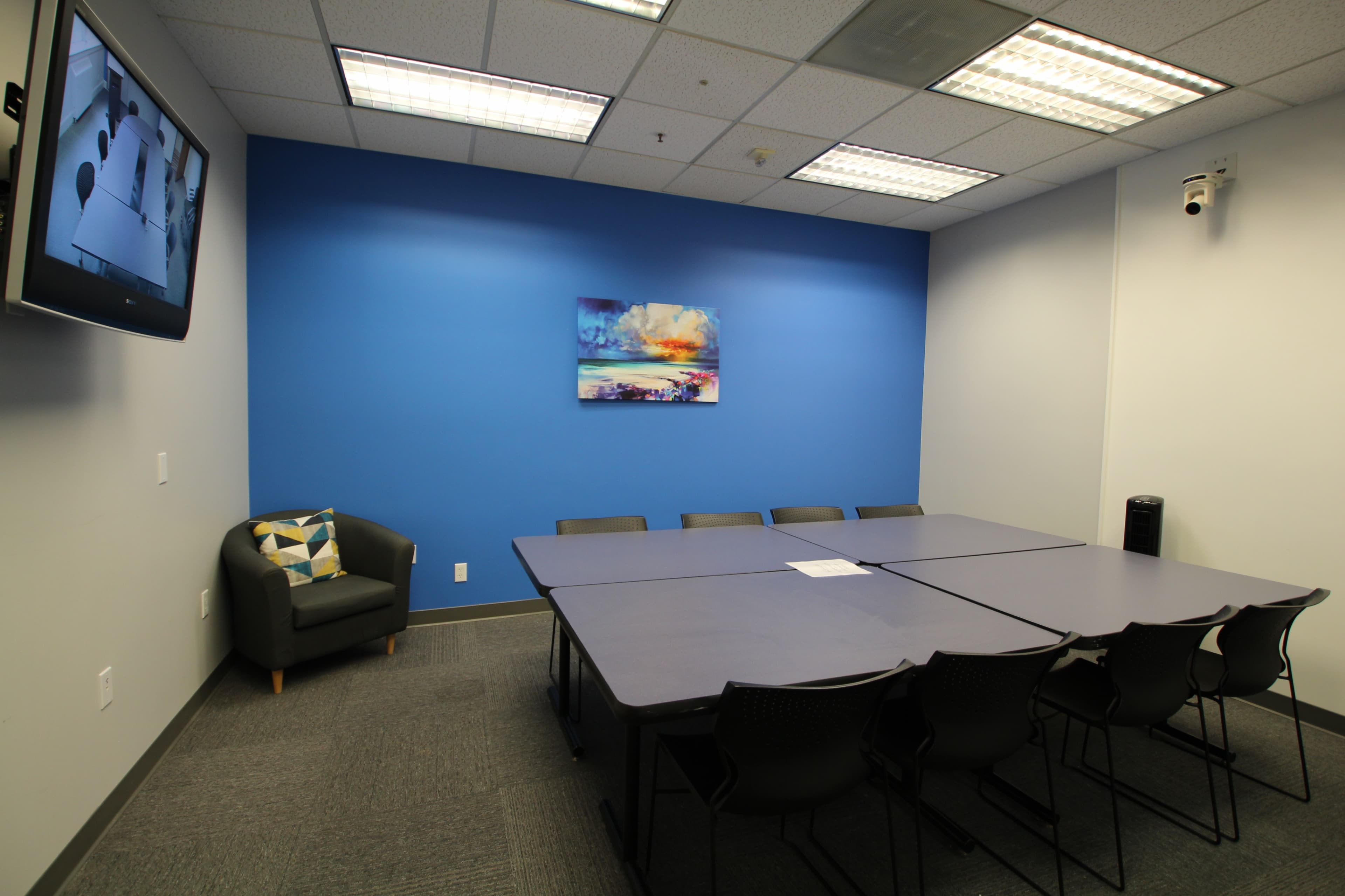 A conference room features a large table surrounded by chairs, with a television and a blue accent wall displaying a landscape painting.