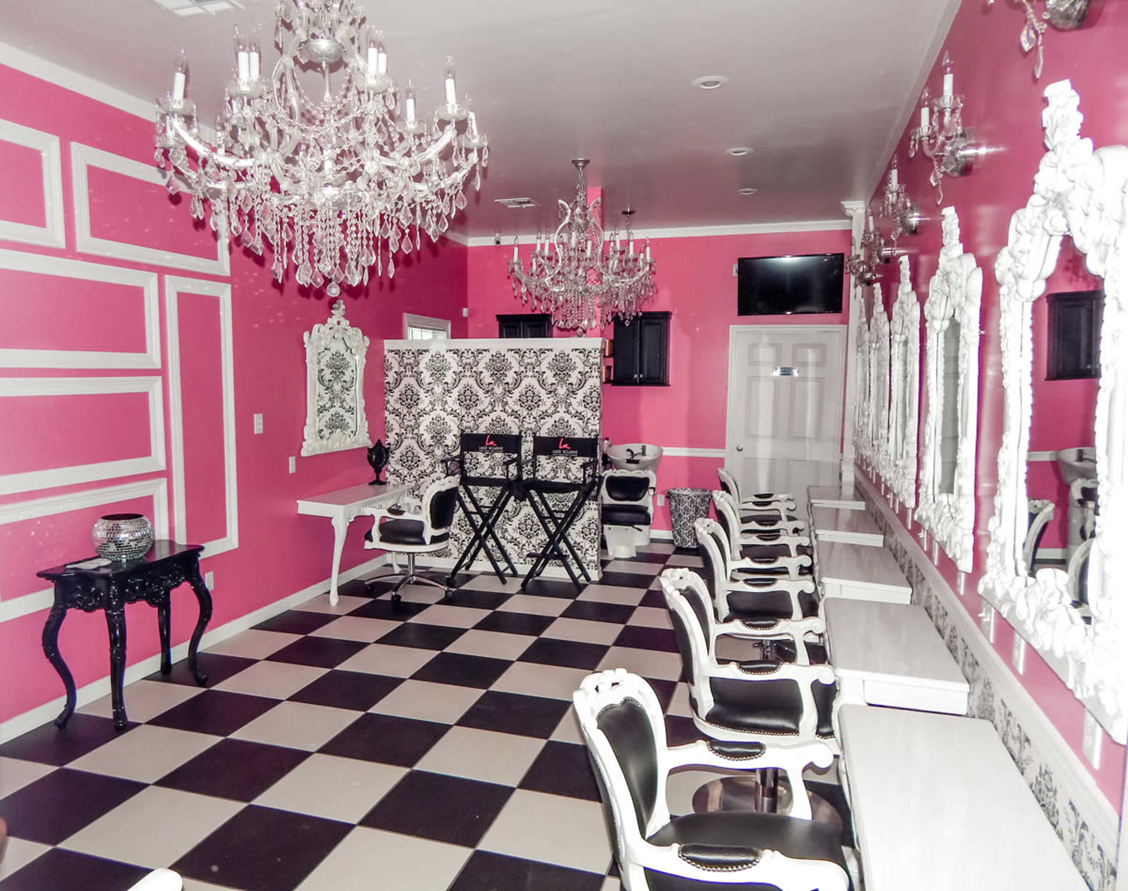 A brightly colored room with pink walls, black and white checkered flooring, elegant chandeliers, and vintage-style furniture arranged around several white tables.