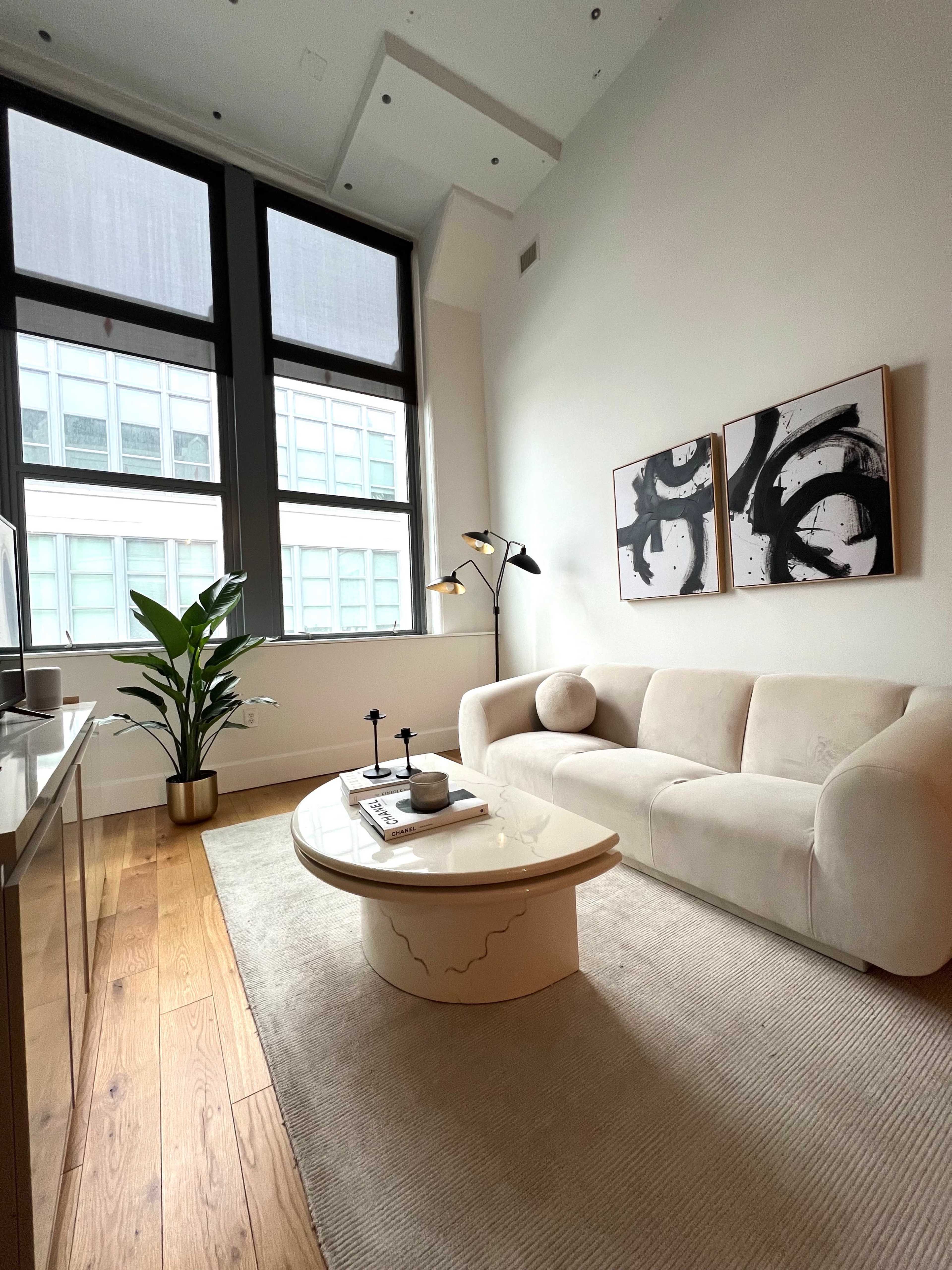 The image shows a modern living room featuring a white sofa, a circular coffee table, a plant, and large windows with a city view.