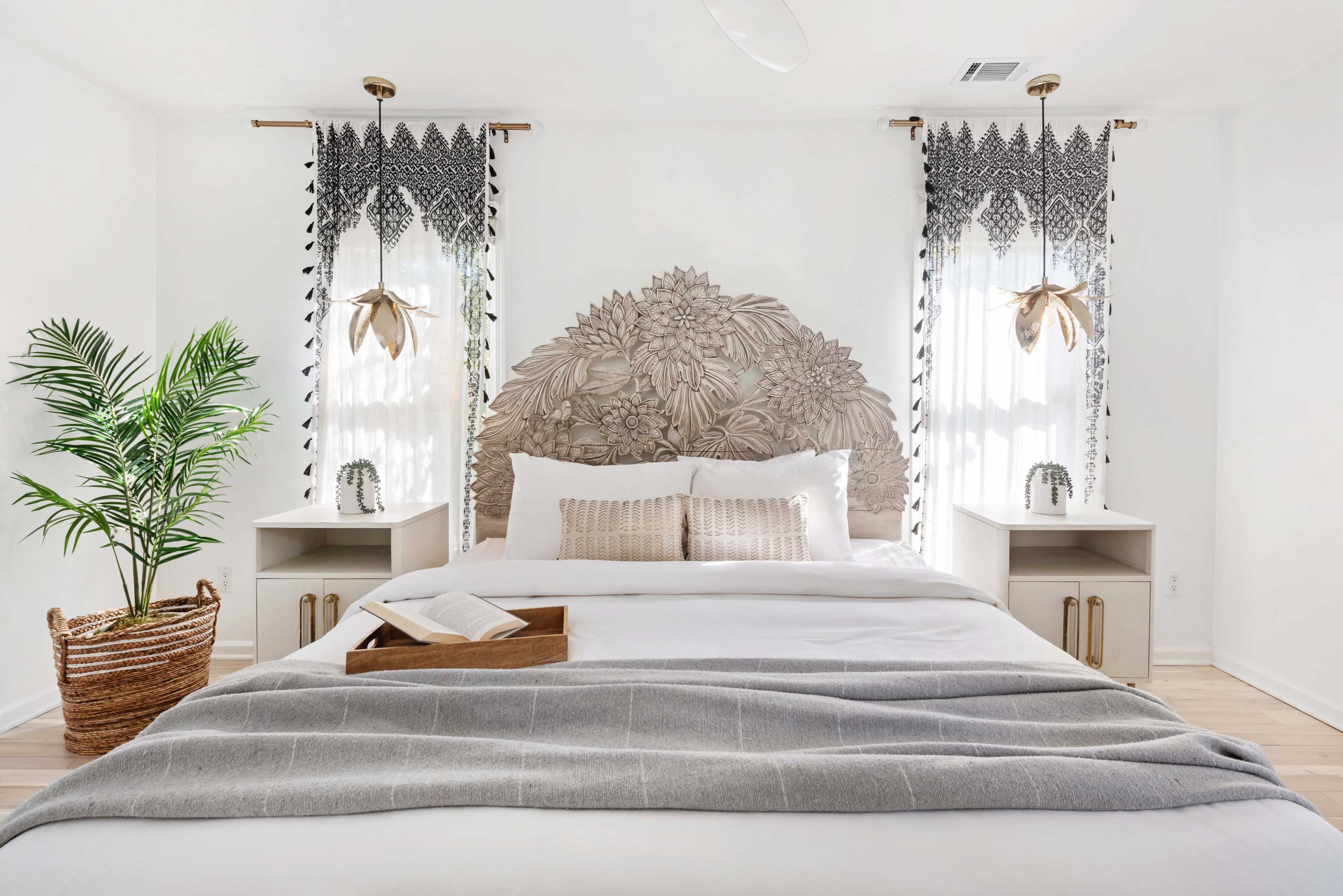 The image shows a neatly arranged bedroom featuring a large, floral headboard, light-colored bedding, and two decorative windows with patterned curtains.