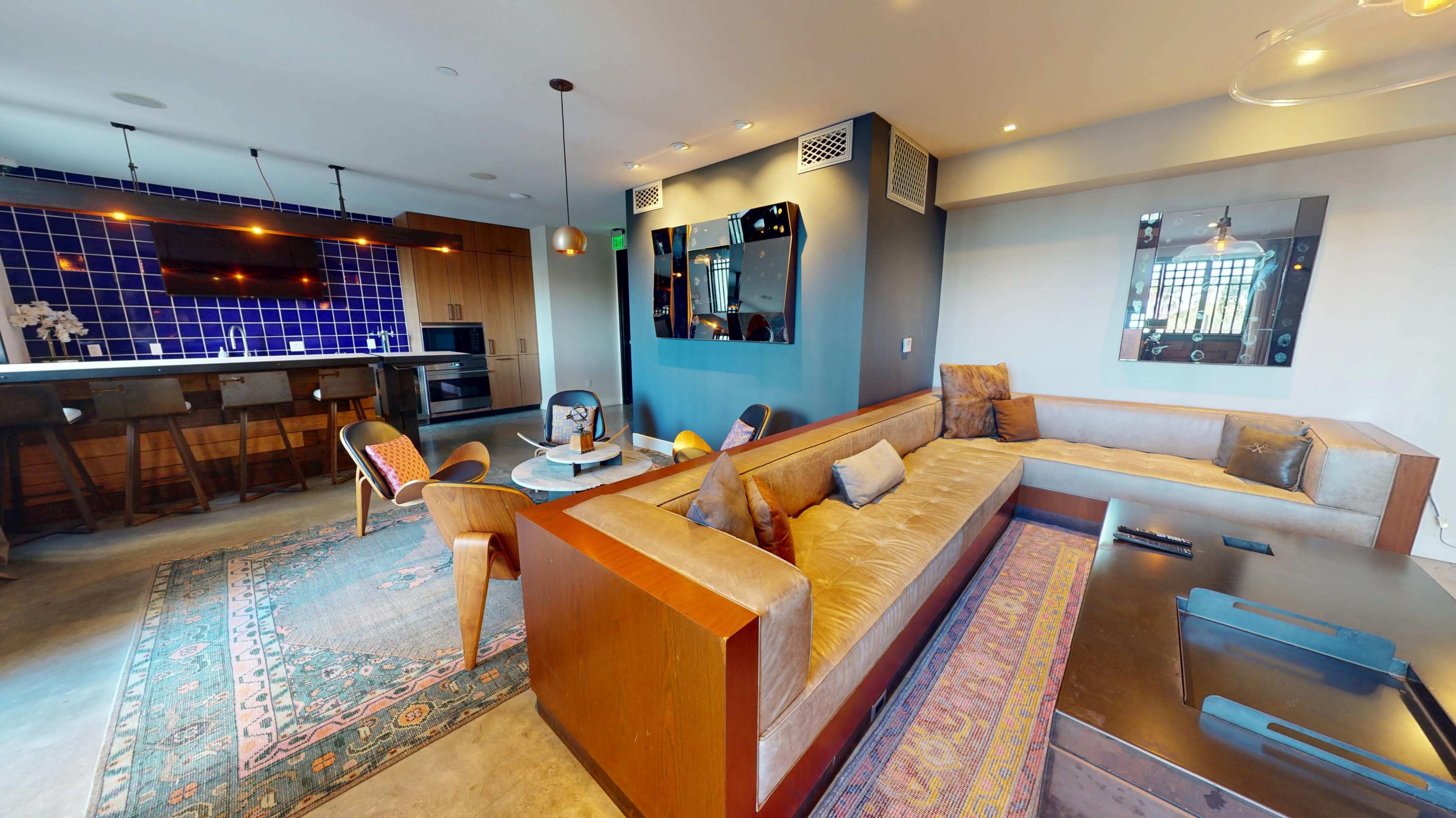 The image shows a modern living space featuring a sectional sofa, a coffee table, and a kitchen area with blue tiled walls and wooden bar seating.